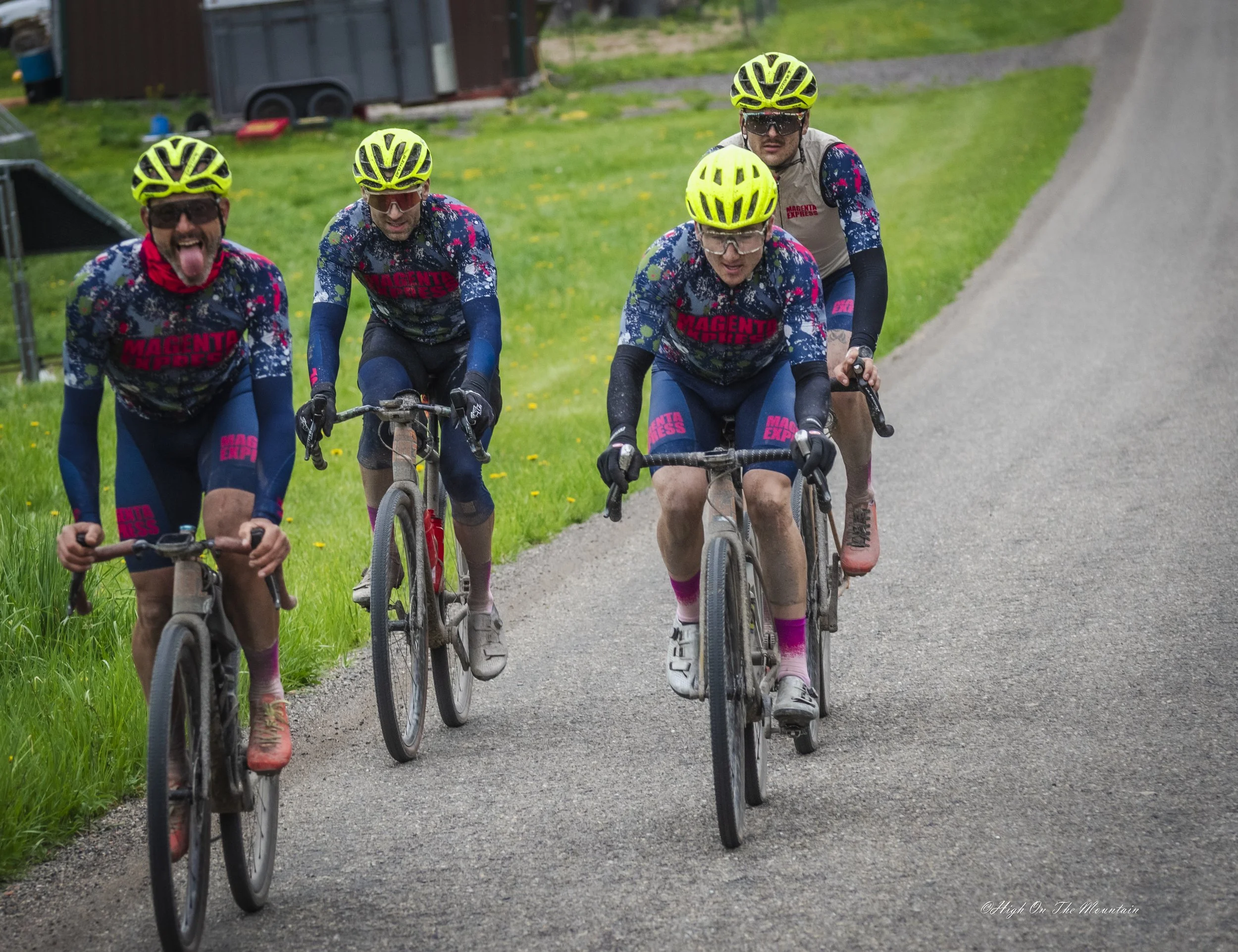 Four cyclists wearing yellow helmets and colorful jerseys riding on a gravel path surrounded by green grass and trees.
