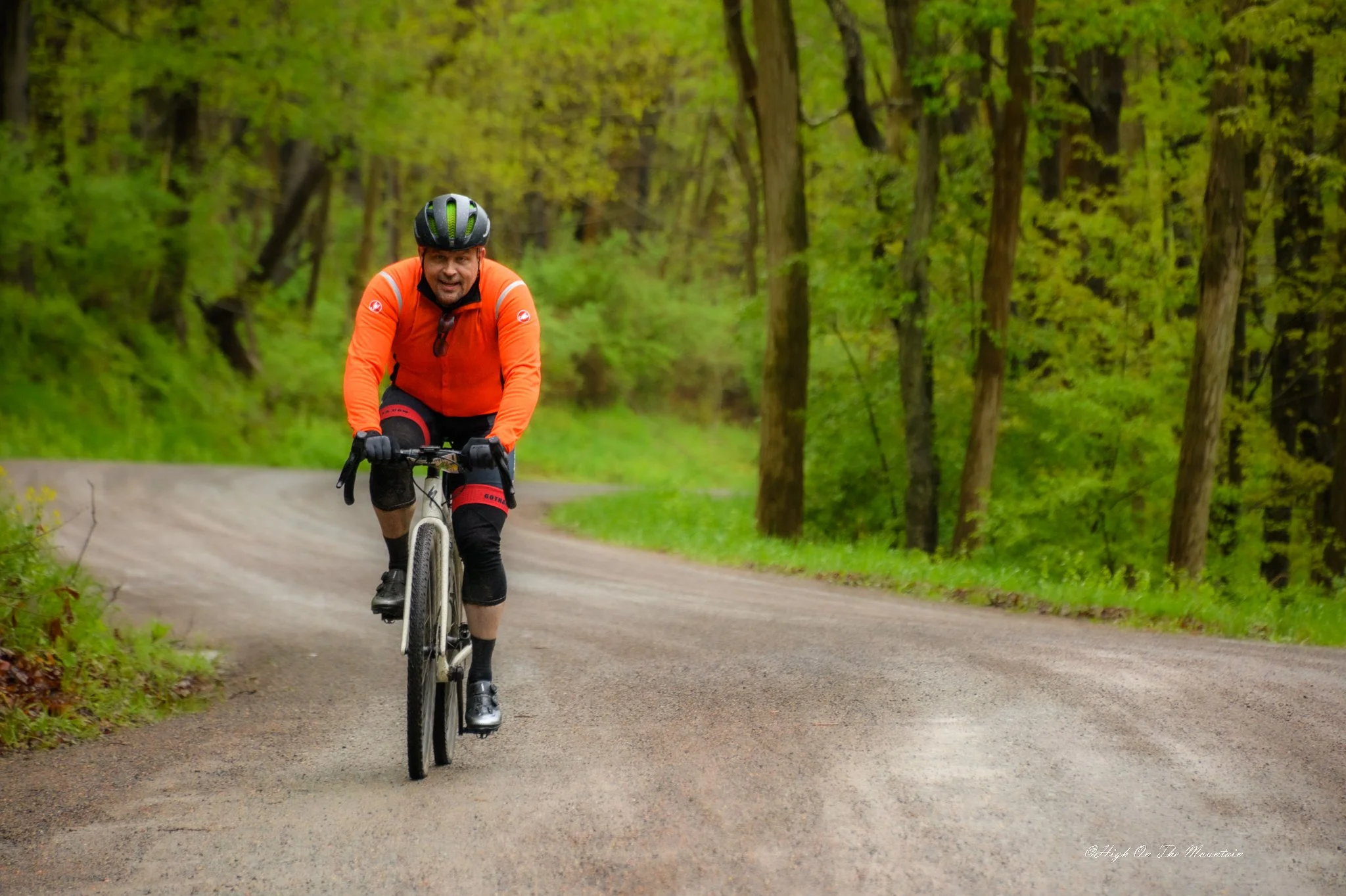 A man wearing a black helmet and orange jacket riding a mountain bike on a dirt trail surrounded by green trees.