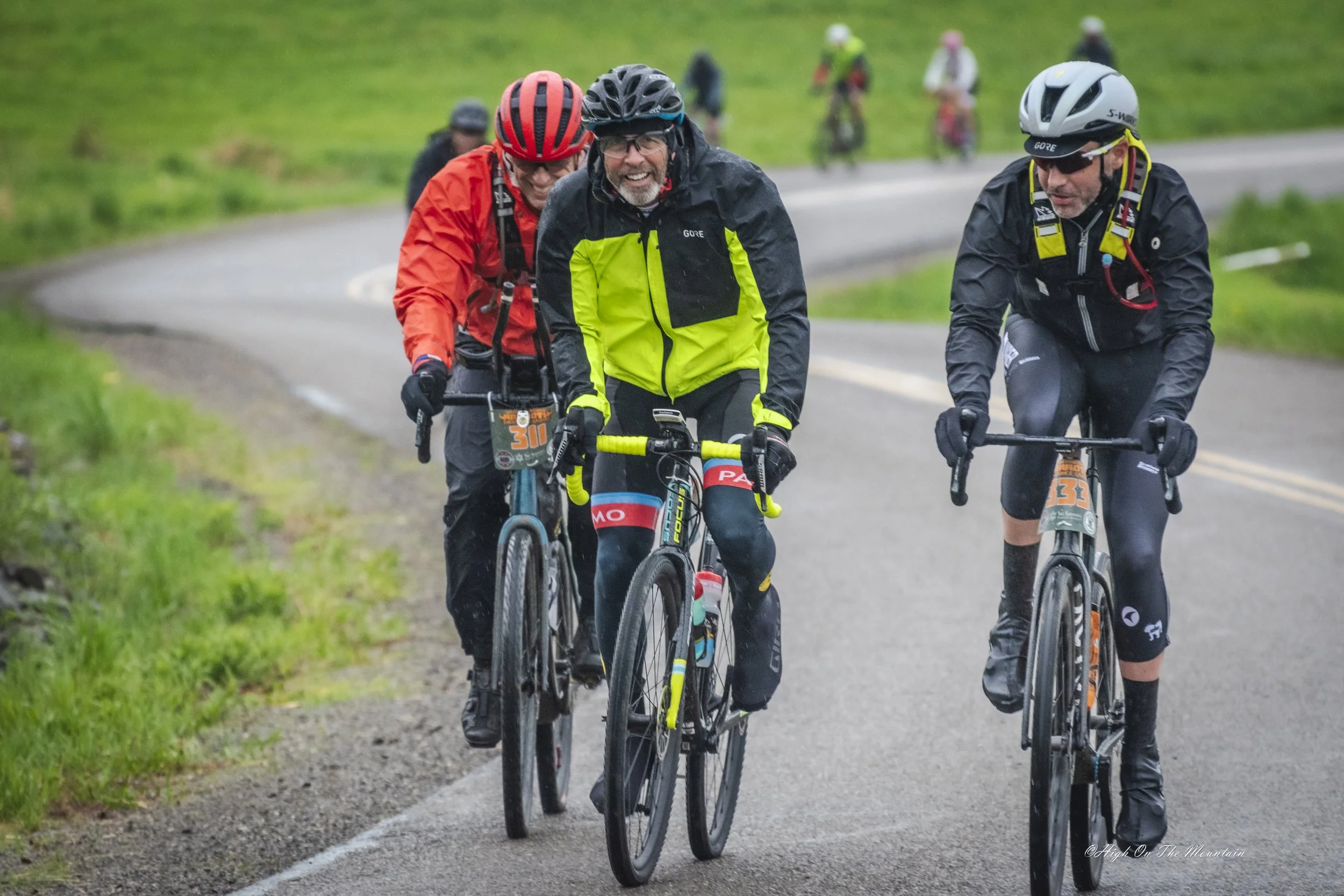 Three cyclists riding on a rural paved road with green grass on the sides, smiling and chatting, all wearing helmets and cycling gear.