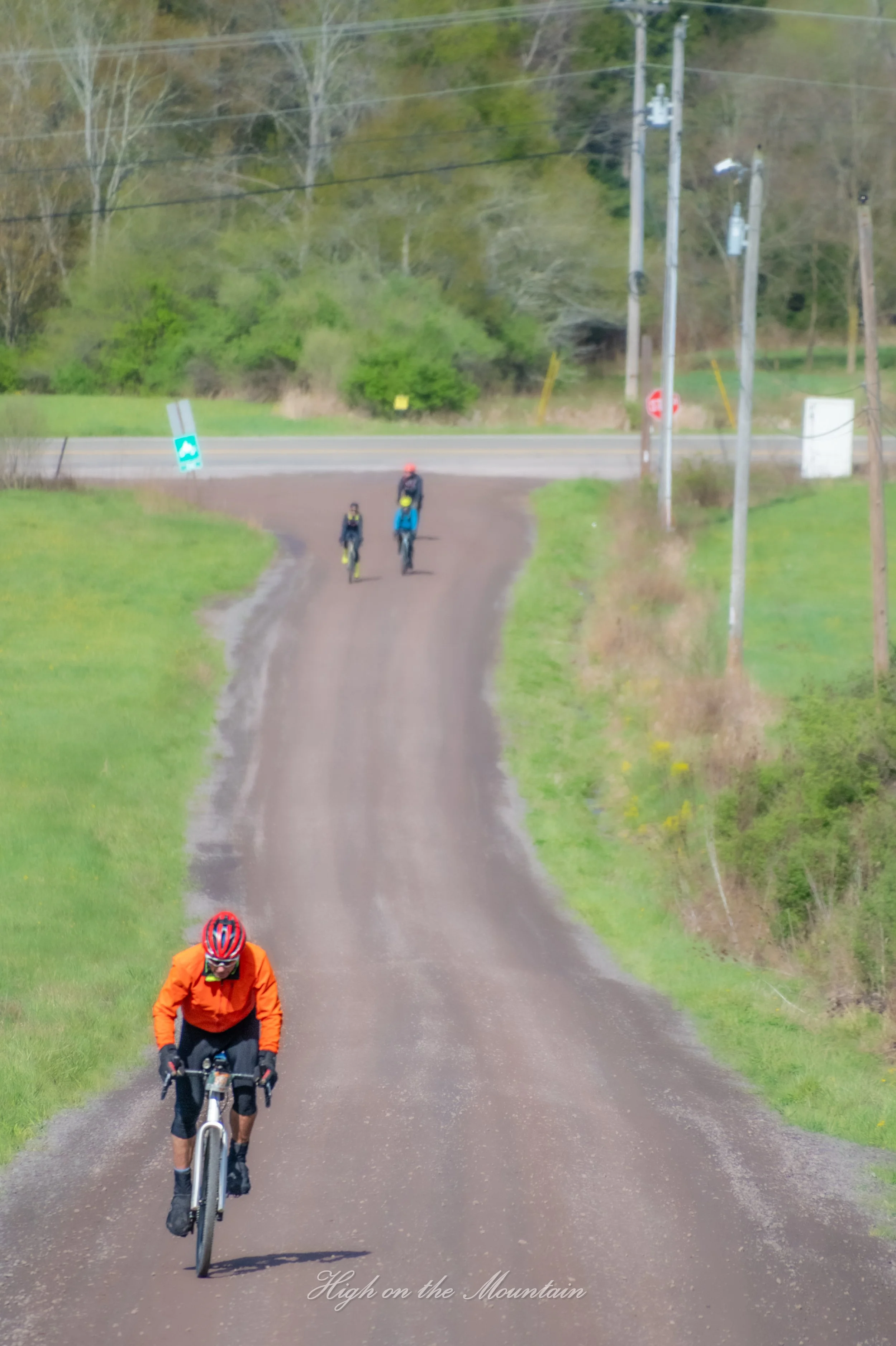Cyclists ride on a dirt trail amidst green grass and trees, with utility poles and a road in the background. The phrase 'High on the Mountain' is written at the bottom of the image.