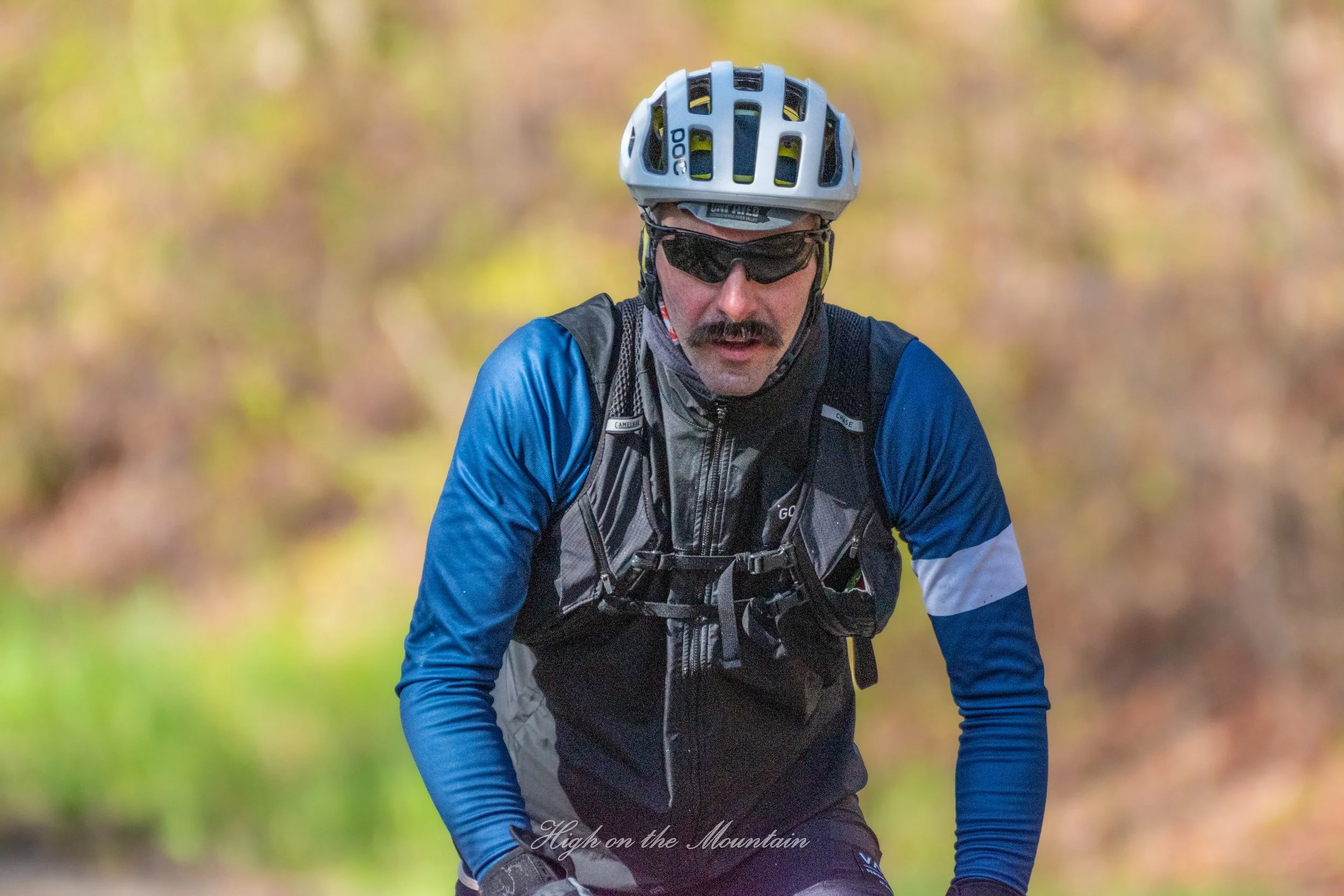 A man mountain biking outdoors wearing a blue jacket, black vest, helmet, and sunglasses, with autumn-colored trees in the background.