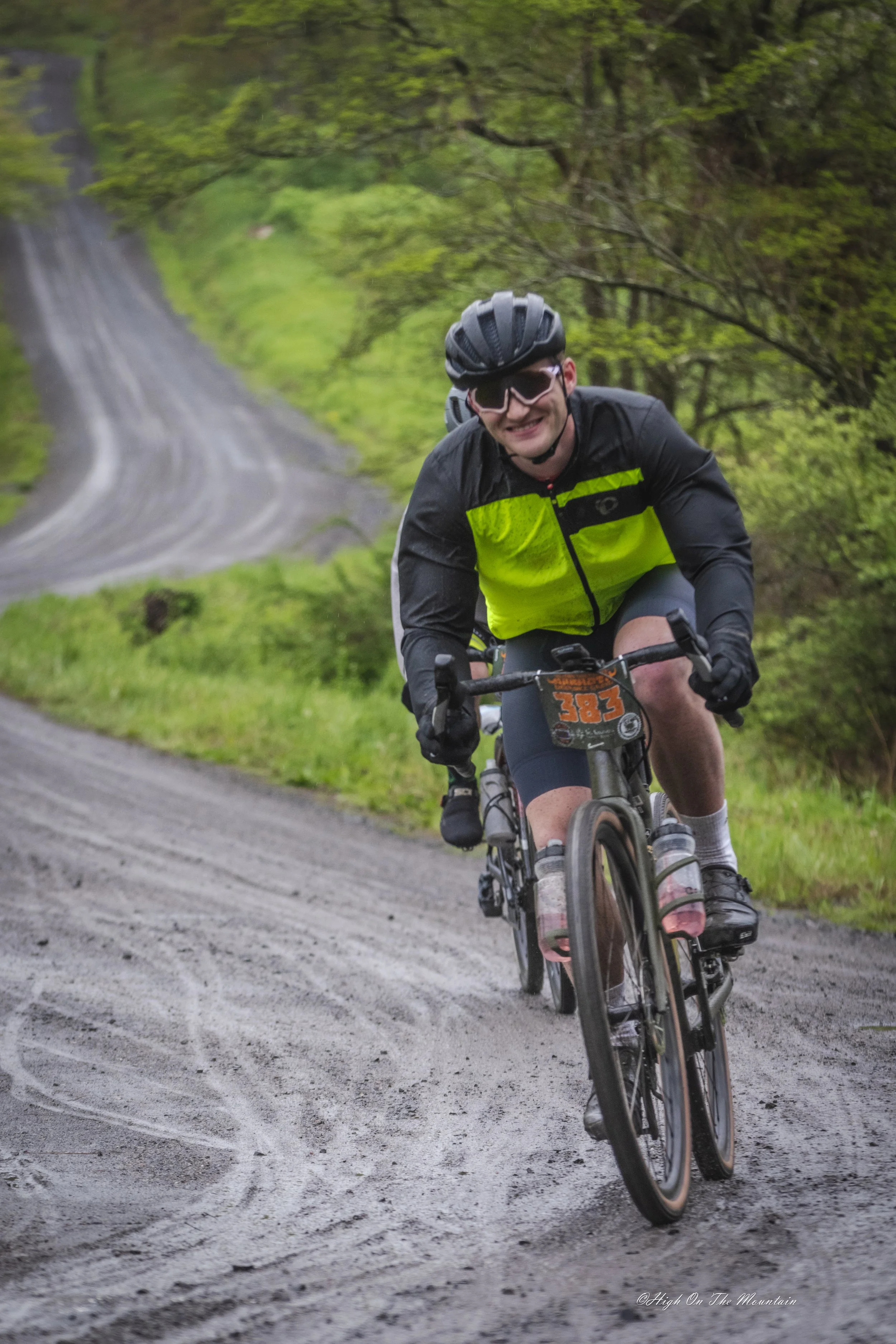 A man riding a mountain bike on a winding dirt trail through a lush green forest. He is wearing a black helmet, sunglasses, a black and neon green jacket, and black shorts. He is smiling and leaning forward as he pedals up a slight incline.