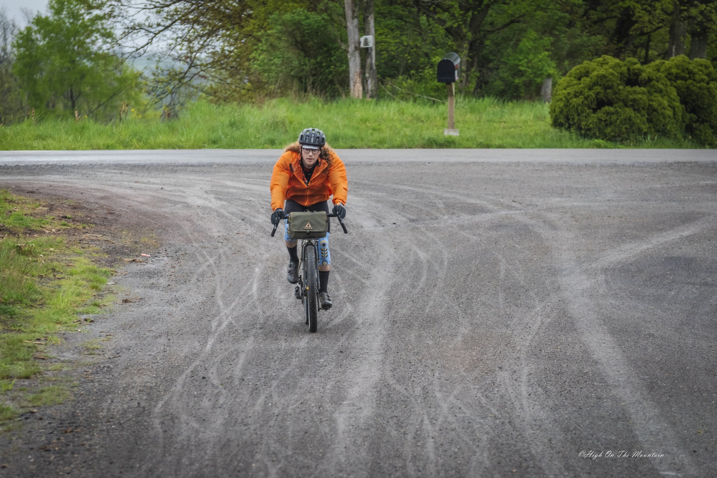 A woman wearing an orange jacket, helmet, and gloves riding a bicycle on a dirt road with green trees in the background.