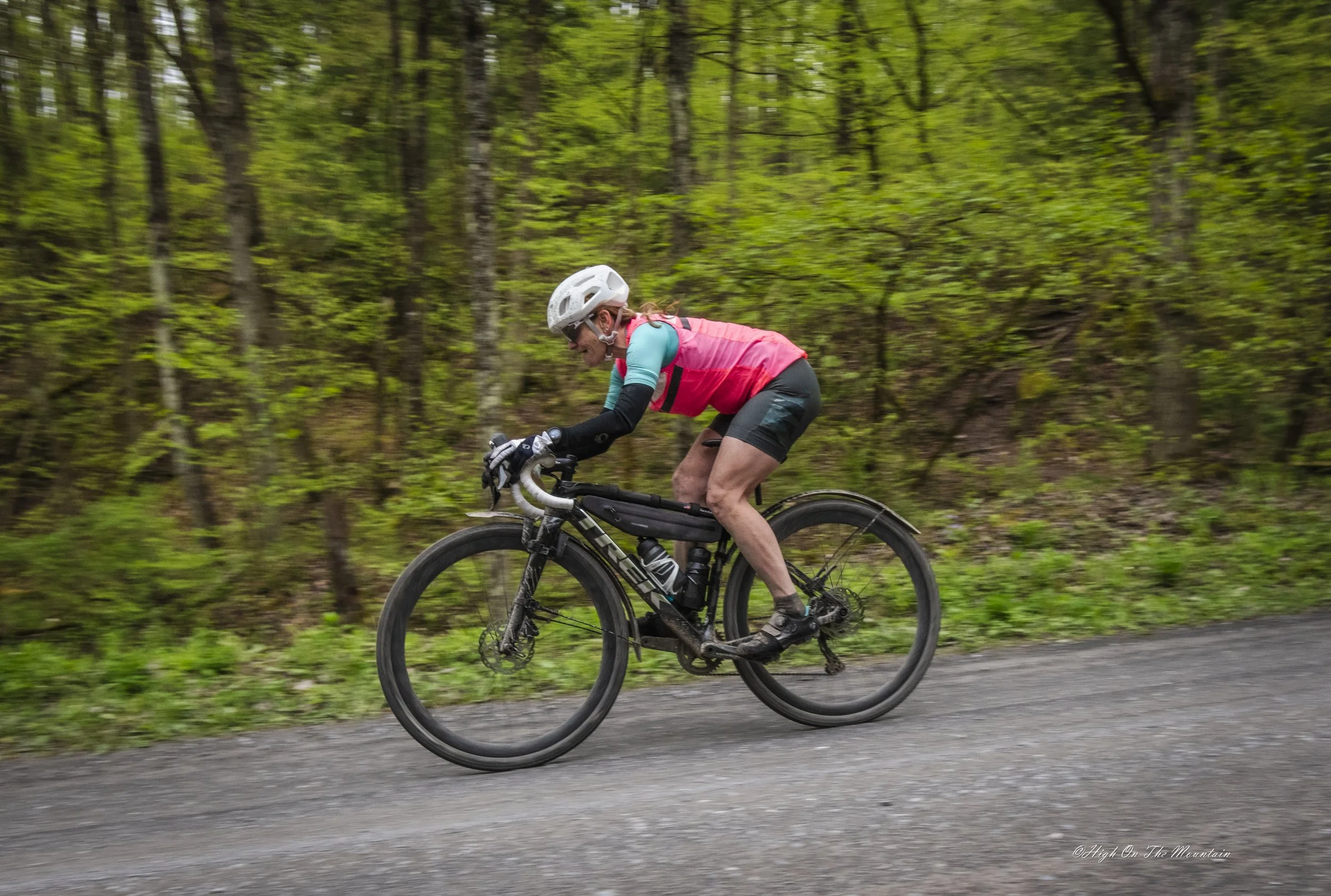 A woman riding a black mountain bike on a gravel trail through a green forest, wearing a white helmet, sunglasses, a pink and teal cycling jersey, black shorts, and gloves.