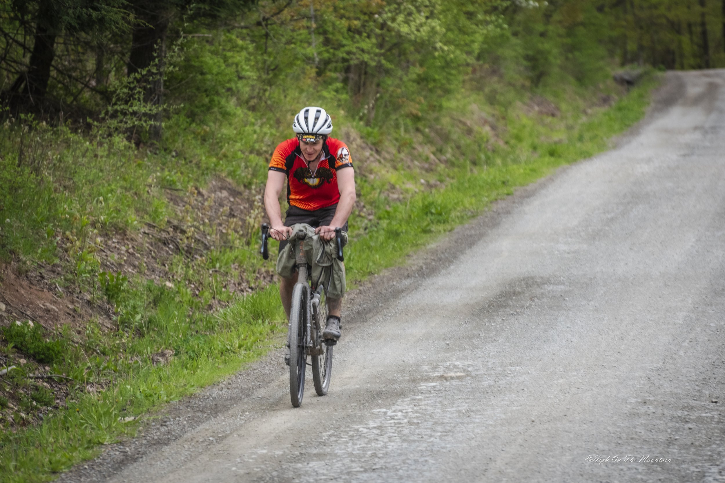A person wearing a red and black cycling jersey, black shorts, and a white helmet is riding a bicycle on a dirt trail surrounded by green trees and grass.