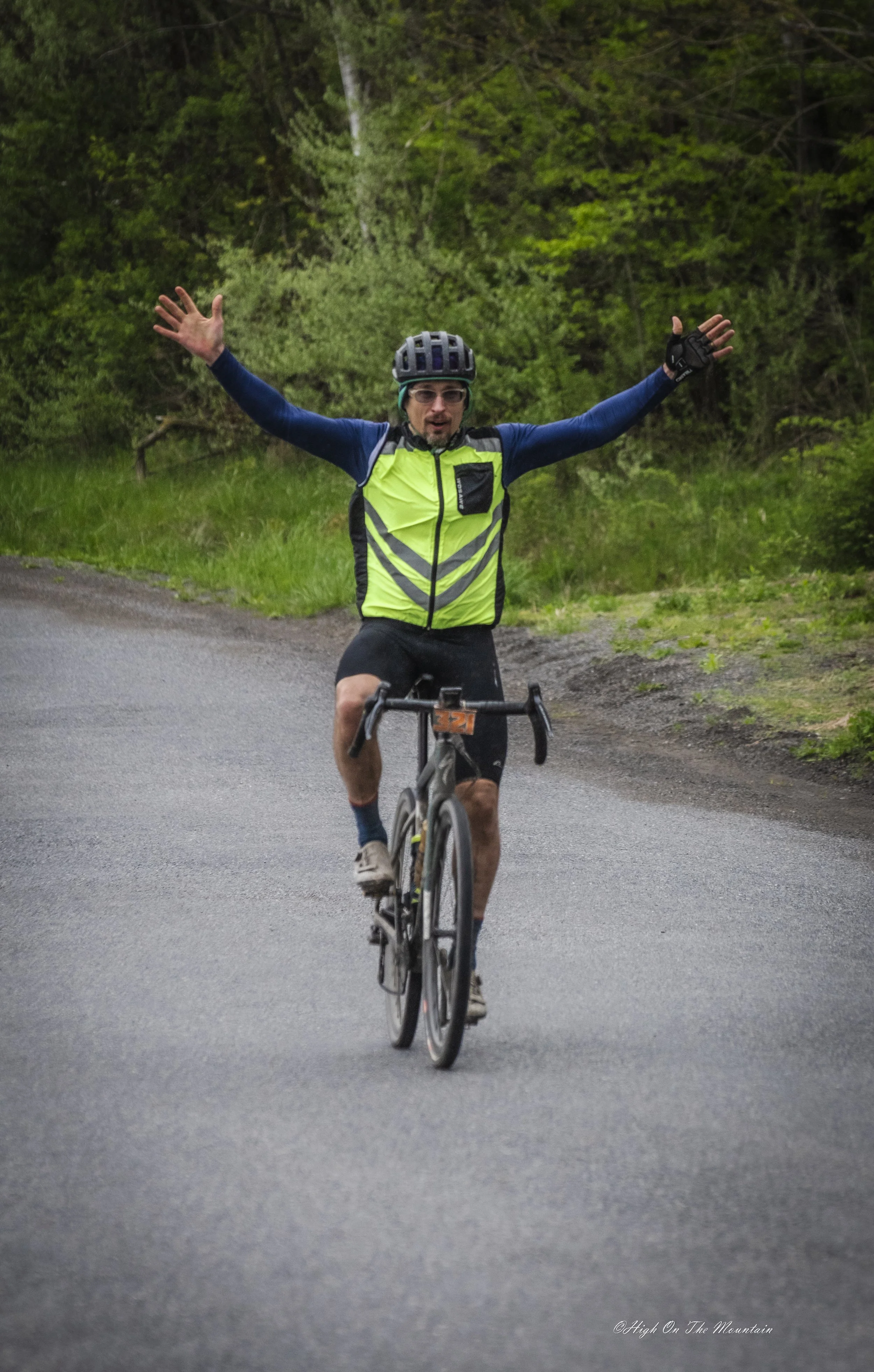A man riding a bicycle on a road surrounded by green trees, wearing a black helmet, glasses, a bright yellow-green vest, and black shorts, with his arms raised in celebration.
