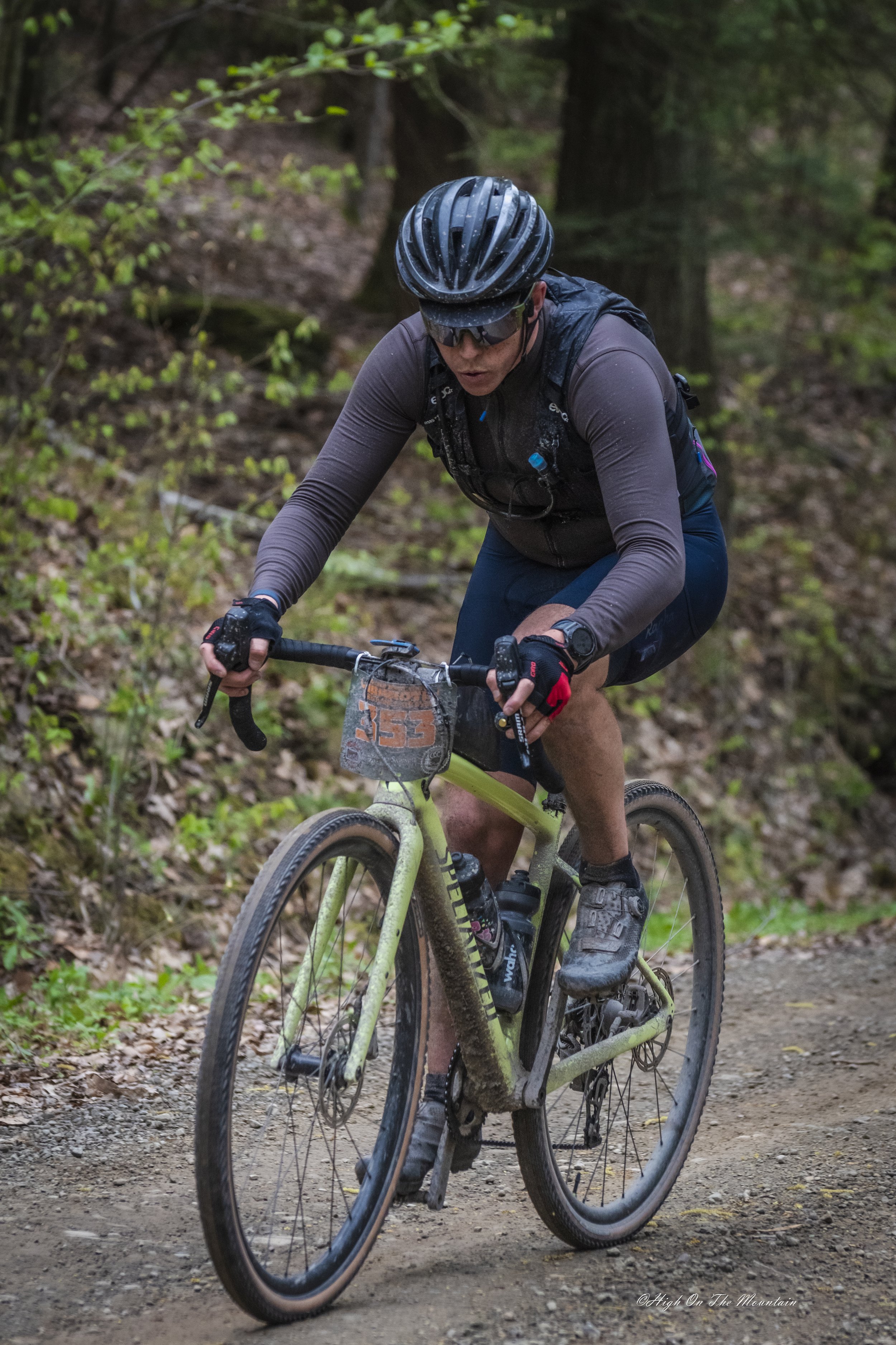 A mountain biker wearing a black helmet, sunglasses, and a dark long-sleeve shirt riding on a dirt trail through a forest with green foliage.