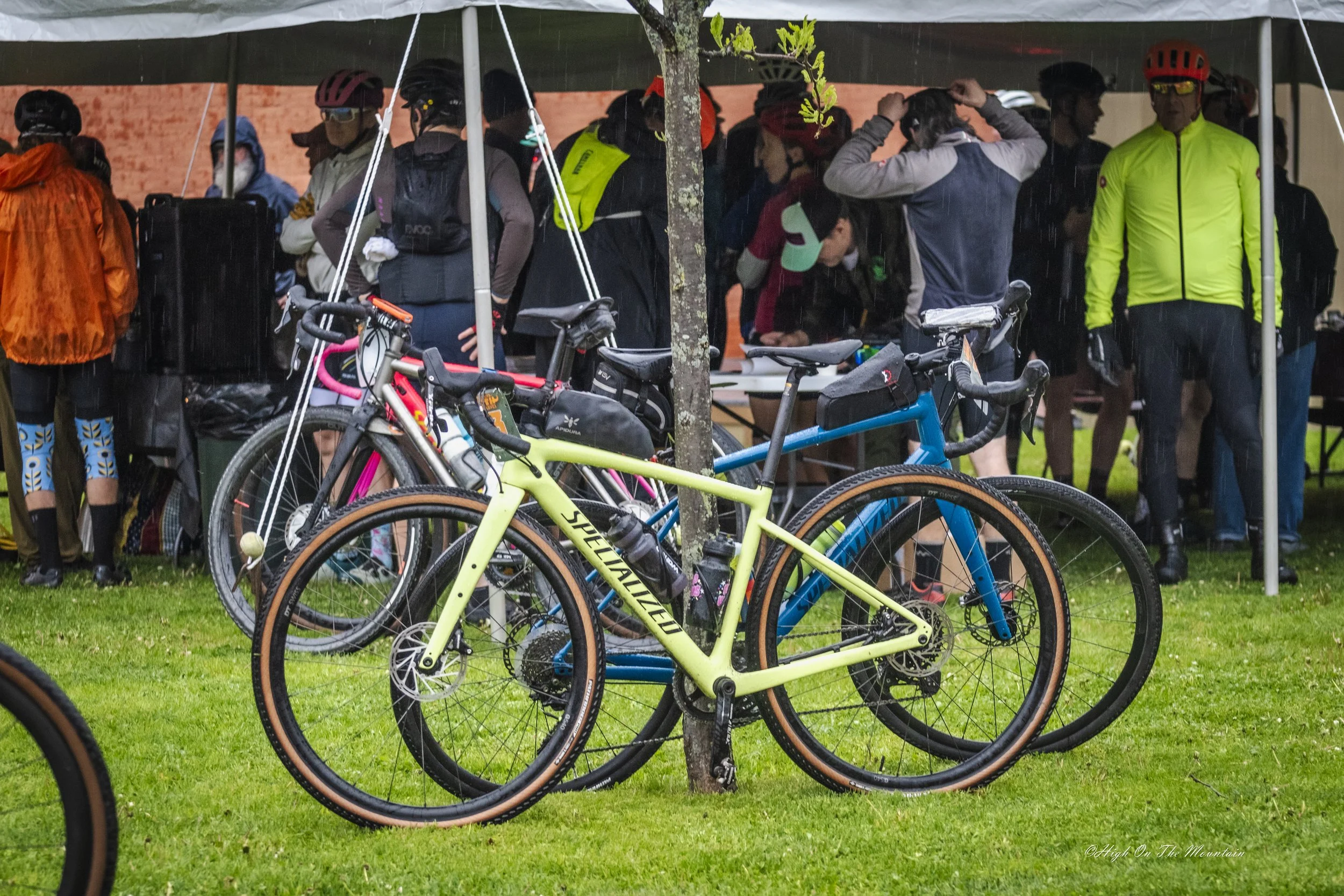 Group of people gathered under a canopy in rain, with bicycles parked in the foreground, some wearing helmets and rain gear.