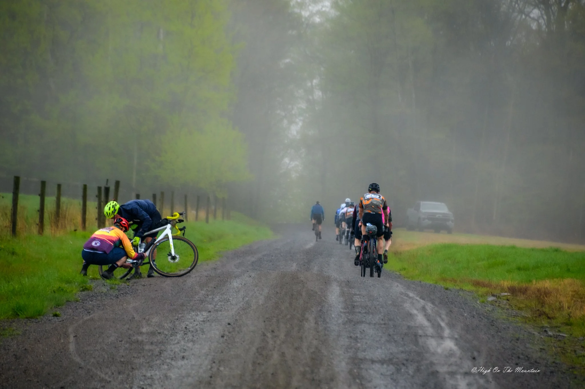 A group of cyclists riding through a foggy, wooded trail on a cloudy day. Some cyclists are stopped and adjusting their bikes, while others ride in the background.