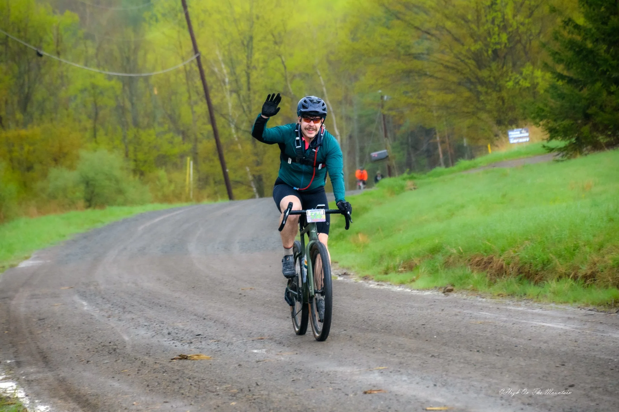 A man wearing a helmet and sunglasses riding a mountain bike on a dirt trail in a green, wooded area, waving at the camera.