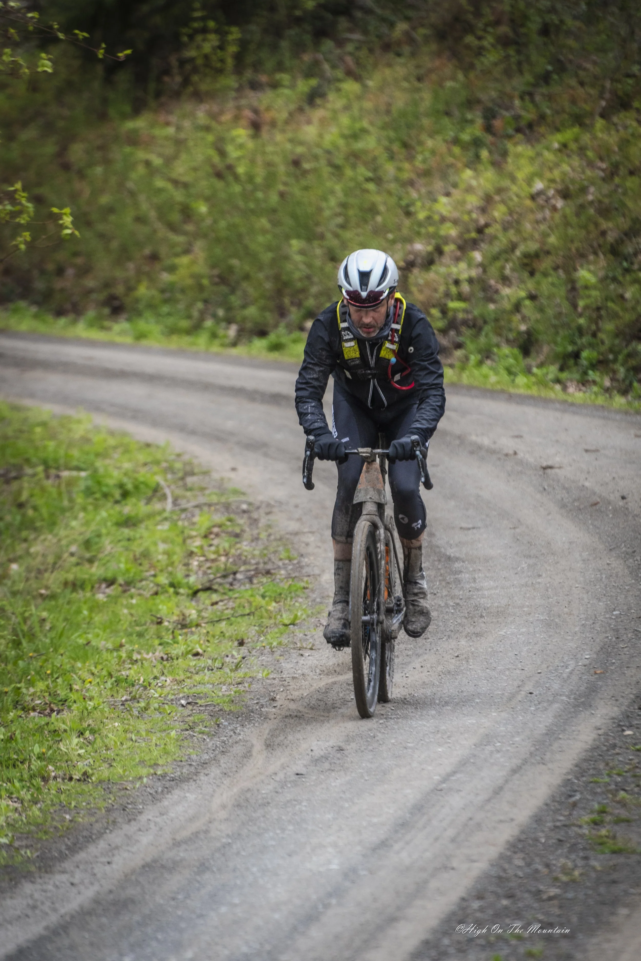 A man wearing a helmet, black jacket, and riding a mountain bike on a dirt trail surrounded by greenery.