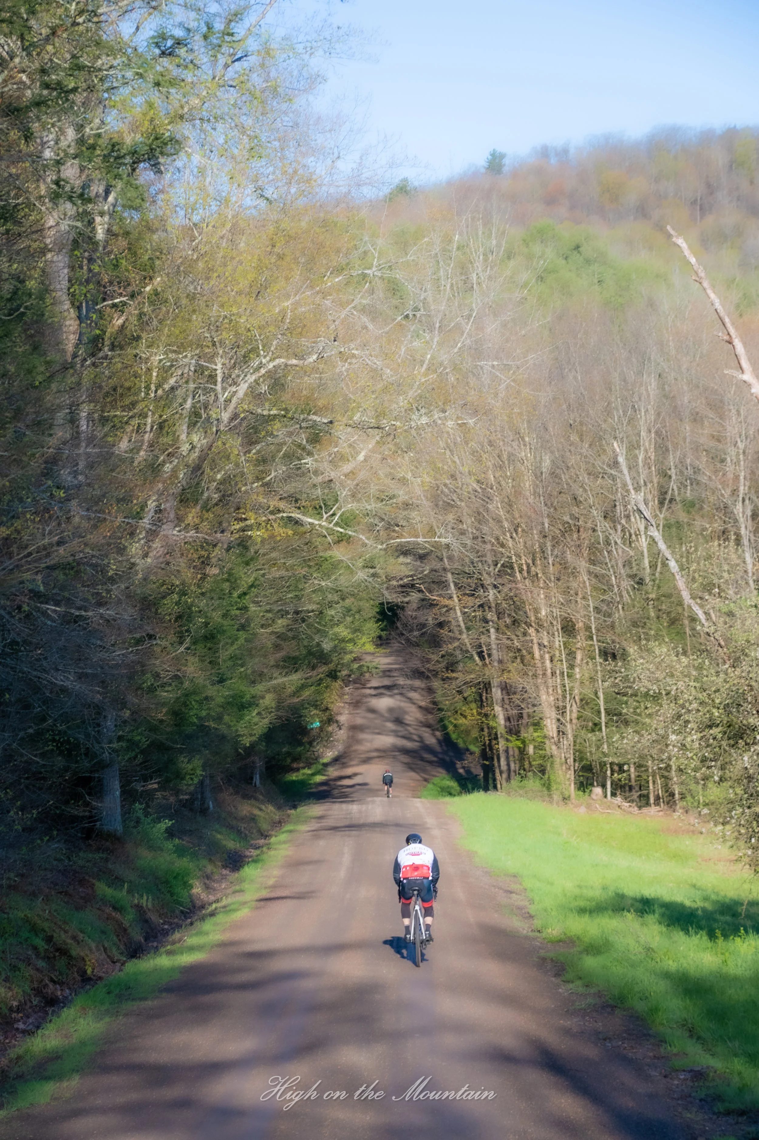 A dirt road running through a wooded area on a mountain, with two cyclists riding on it. The trees are mostly bare and the sky is clear and blue. The text "High on the Mountain" is written in cursive at the bottom of the image.