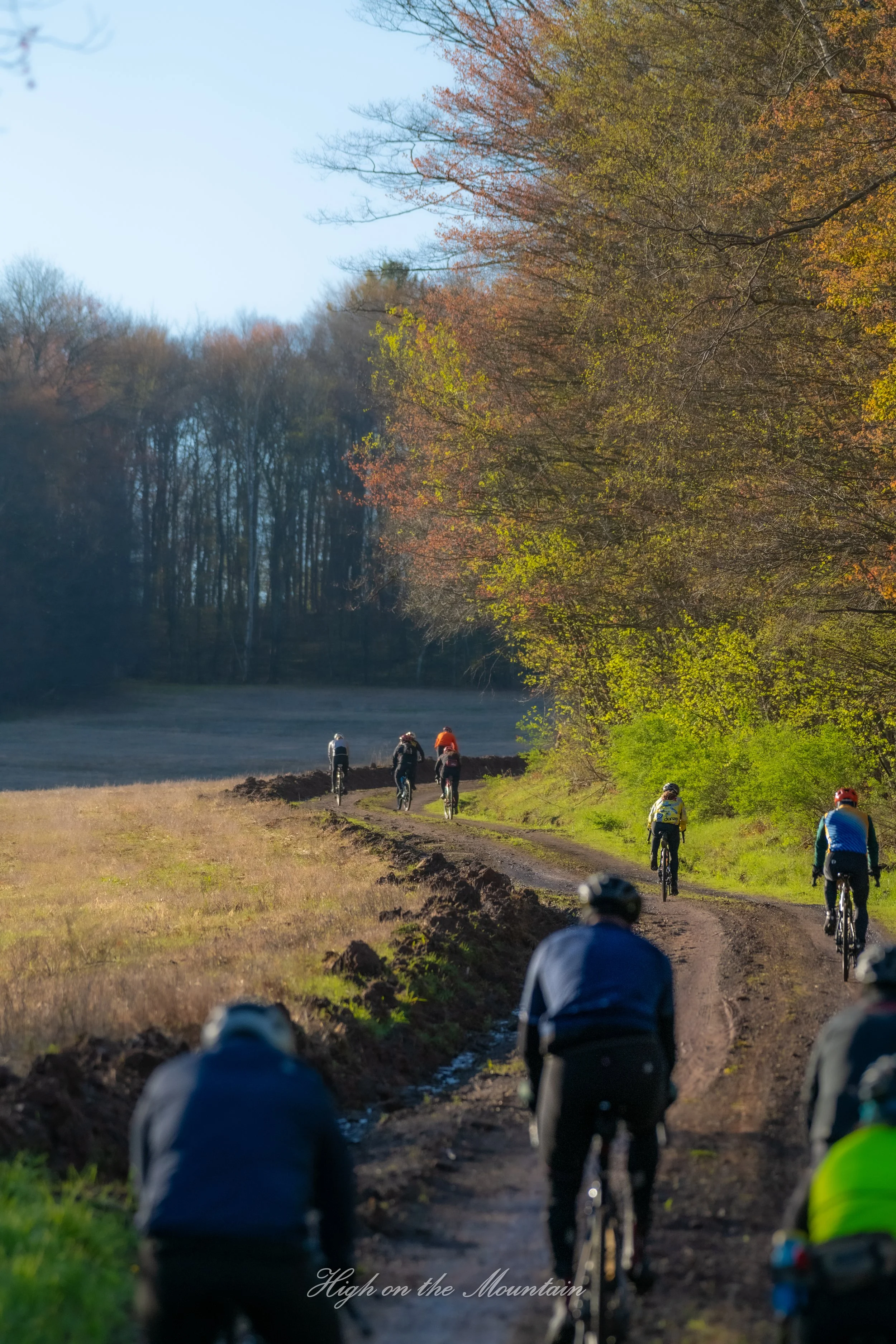 A group of cyclists riding on a dirt trail through a wooded area with trees showing fall foliage. The sky is clear and the background features a forested landscape. The text "High on the Mountain" appears at the bottom of the image.
