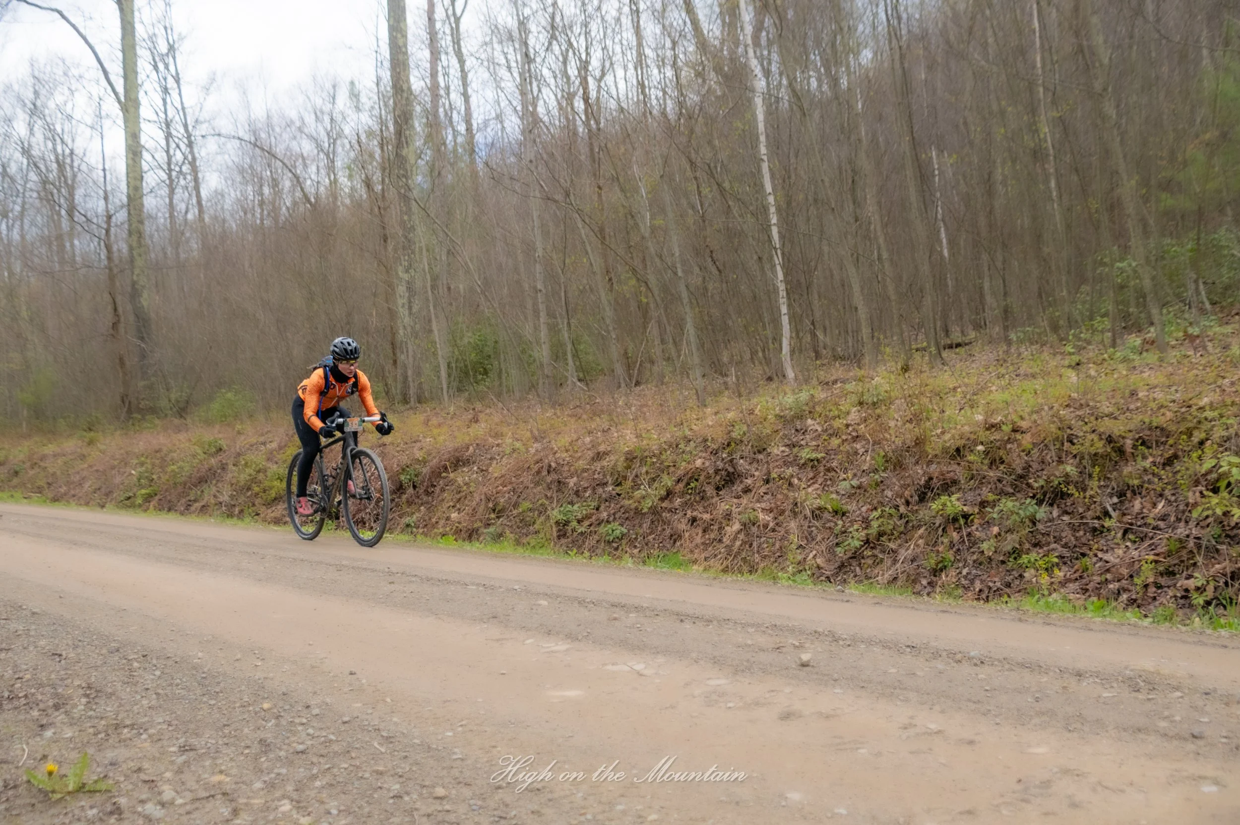 A person wearing orange jacket, helmet, and sunglasses riding a mountain bike on a dirt trail in a forested area with leafless trees.