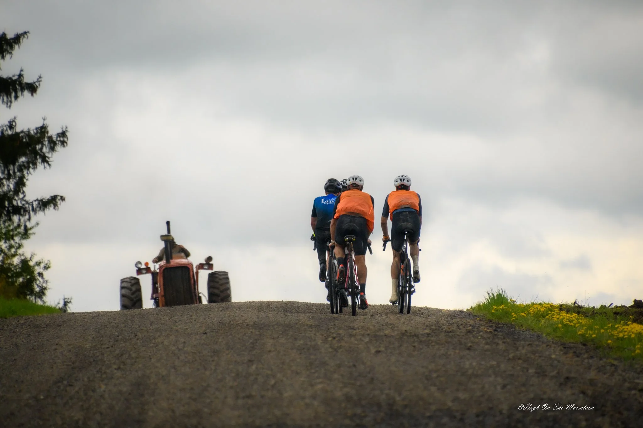 Three cyclists riding uphill on a dirt path under cloudy sky, with a tractor behind them and trees on the side.