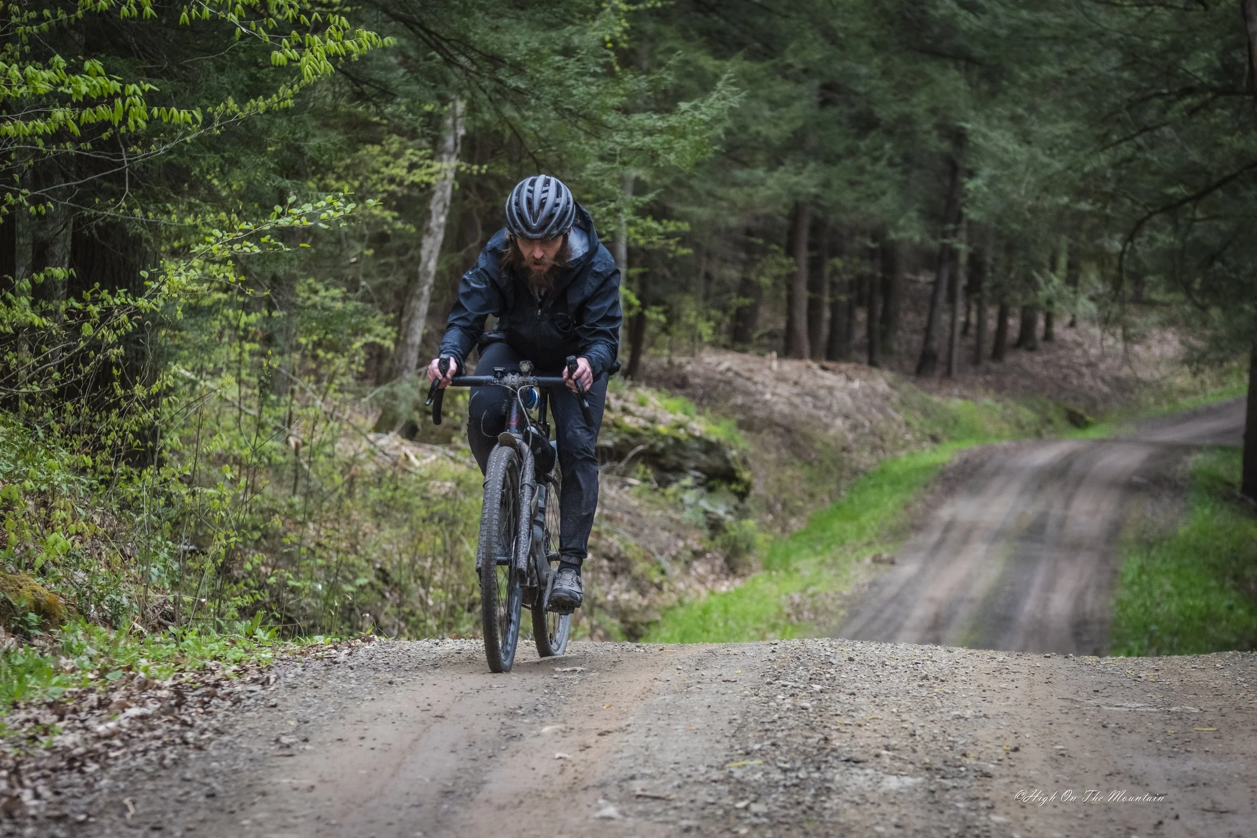 A man riding a mountain bike on a dirt path through a lush, green forest, wearing a helmet and dark outdoor clothing.