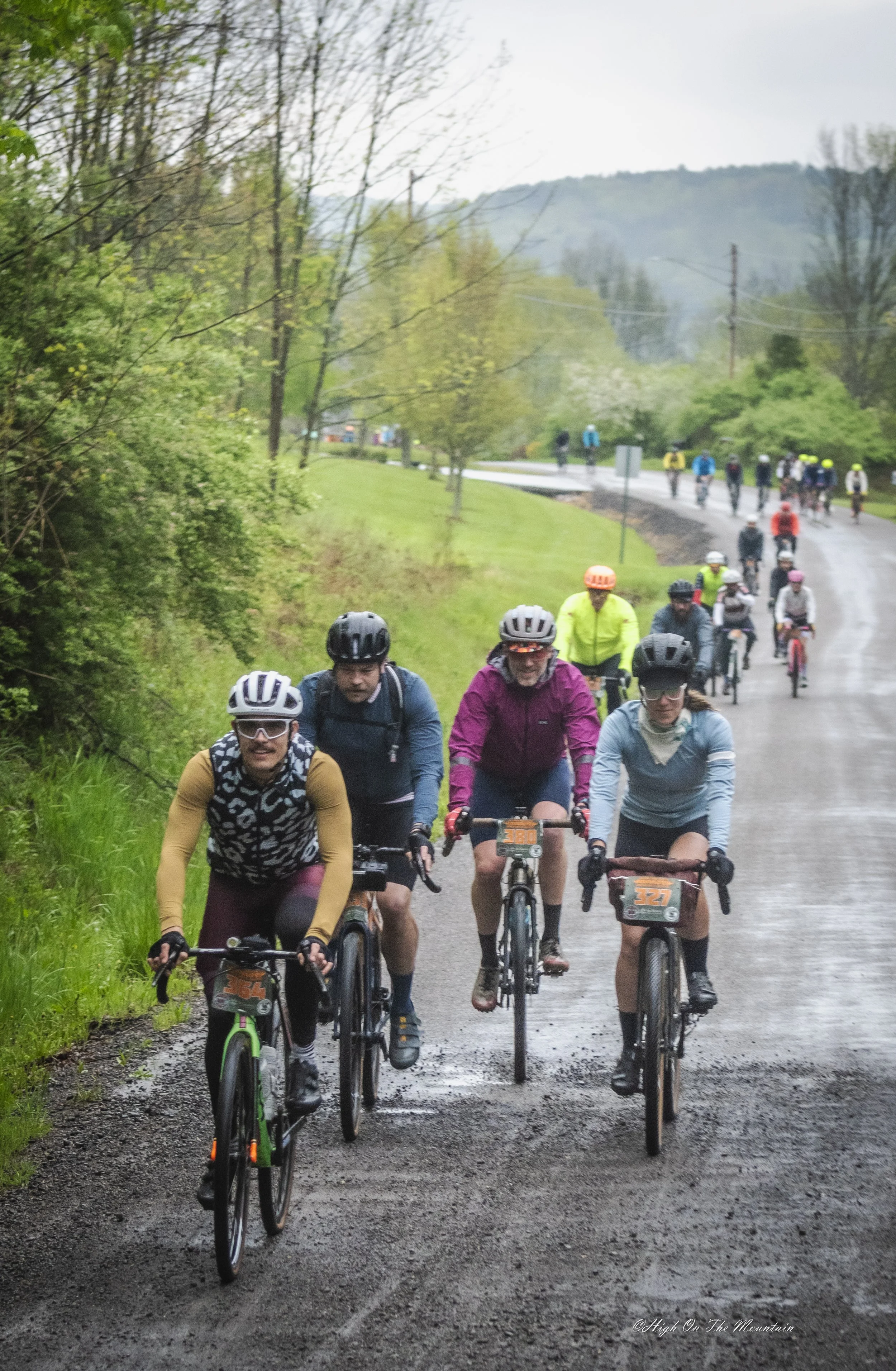 Group of people riding mountain bikes on a dirt trail through a lush, green landscape with trees and hills in the background. The weather appears to be rainy or damp.