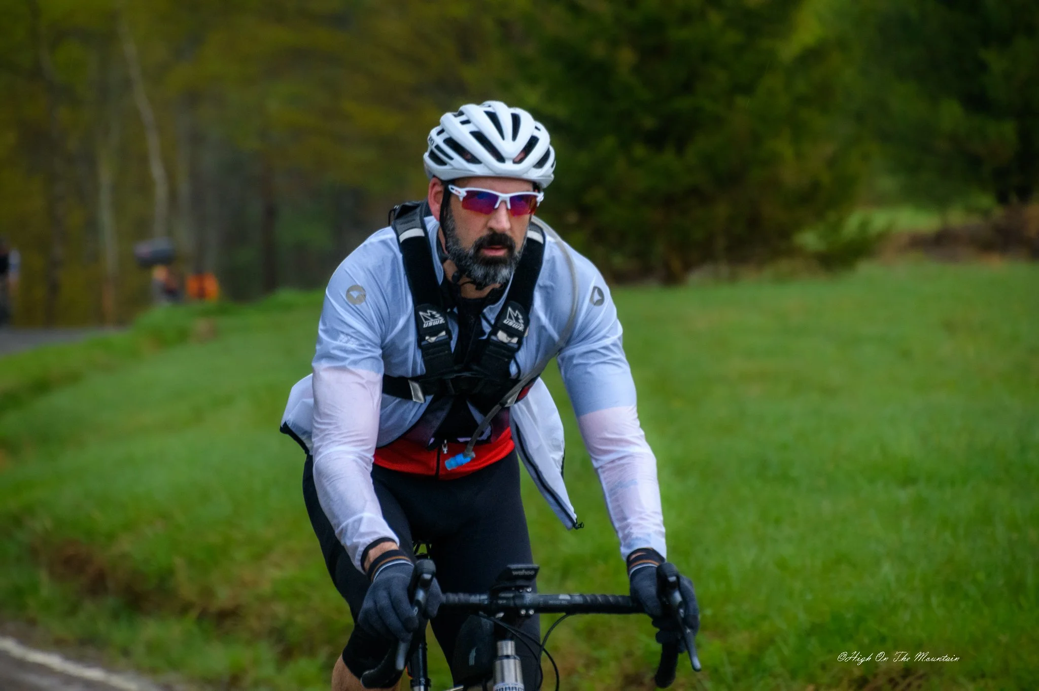 A man with a beard wearing a white bike helmet, sunglasses, and athletic clothing rides a mountain bike along a trail surrounded by green trees.