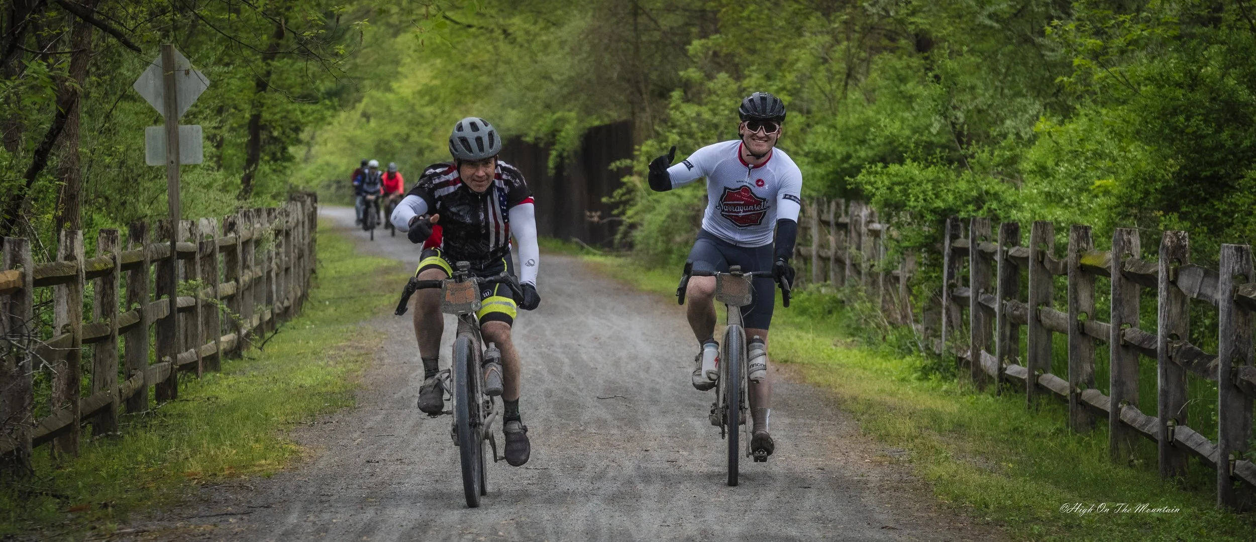Two men riding bicycles on a dirt path through a green forest, smiling and waving, with other cyclists visible in the background.