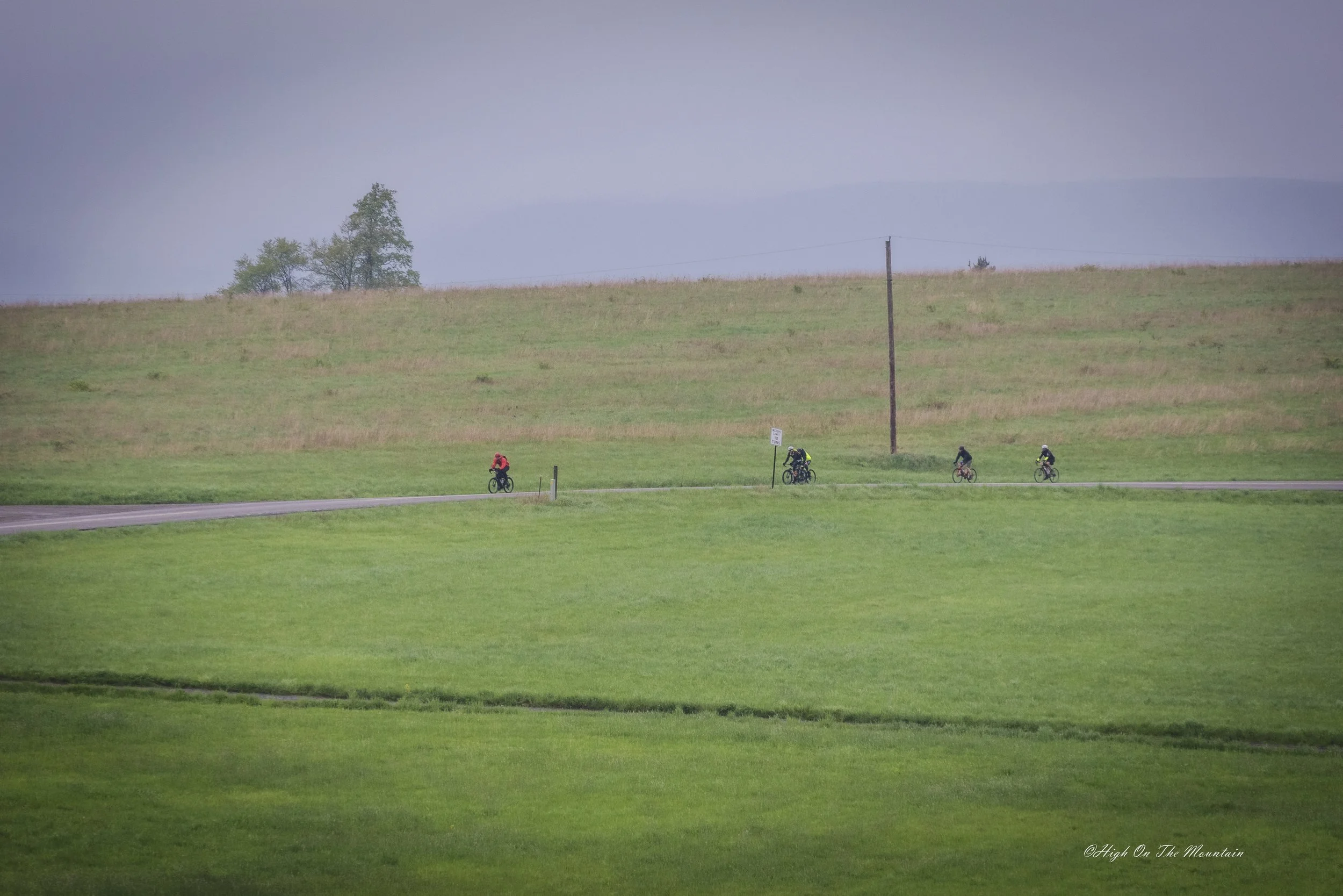 Four cyclists riding along a paved road through lush green fields under a cloudy sky, with a few trees in the distance.