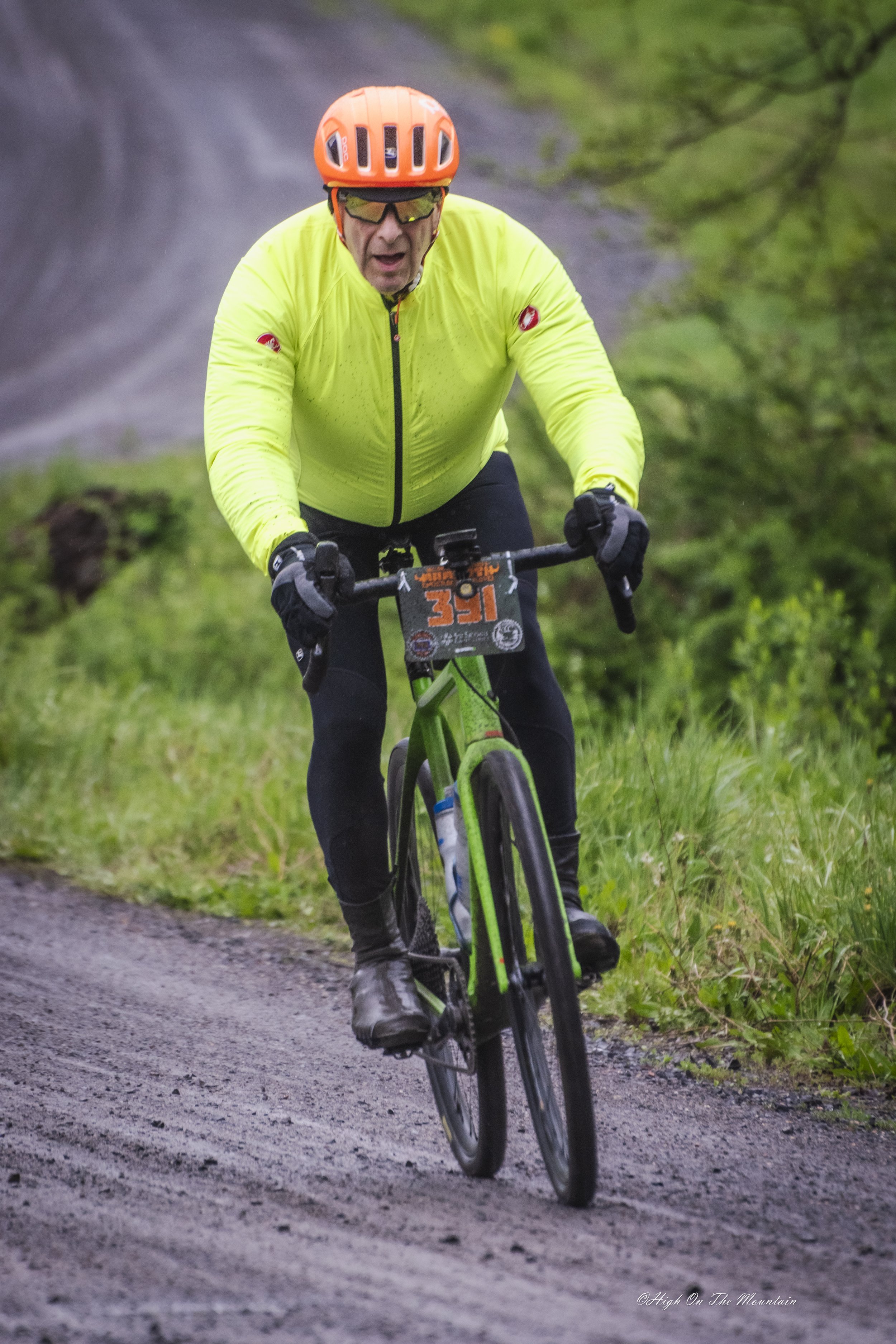 A man wearing a bright yellow jacket, black gloves, orange helmet, and sunglasses riding a green mountain bike on a dirt trail surrounded by greenery and trees.