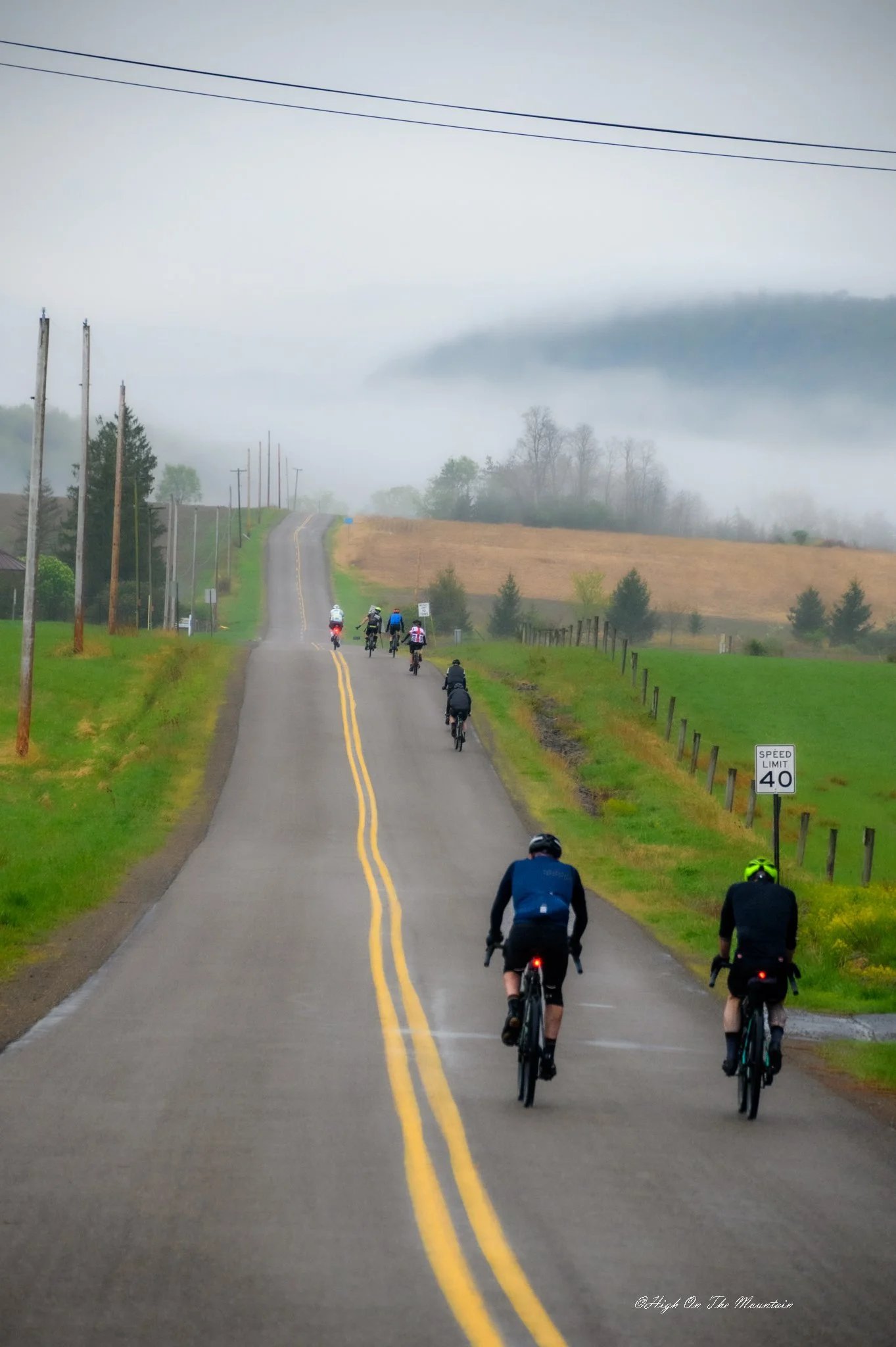 Group of cyclists riding on a misty rural road with green fields and power lines, fog-covered hills in the background, and a speed limit sign showing 40 miles per hour.