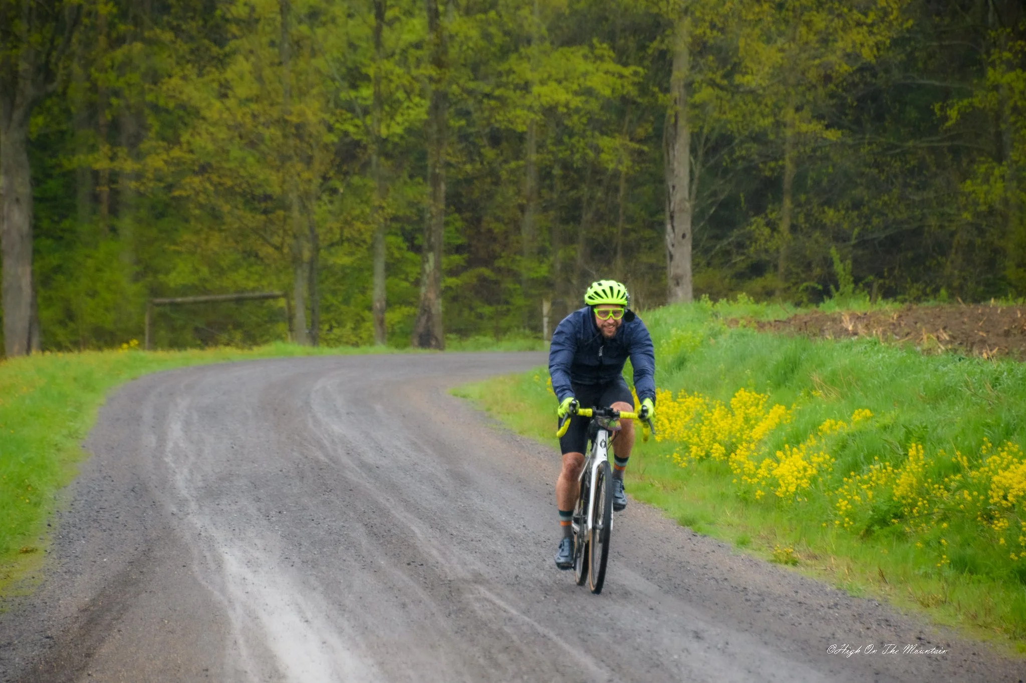 A man riding a bike on a winding gravel road through a green forest with yellow wildflowers on the side.