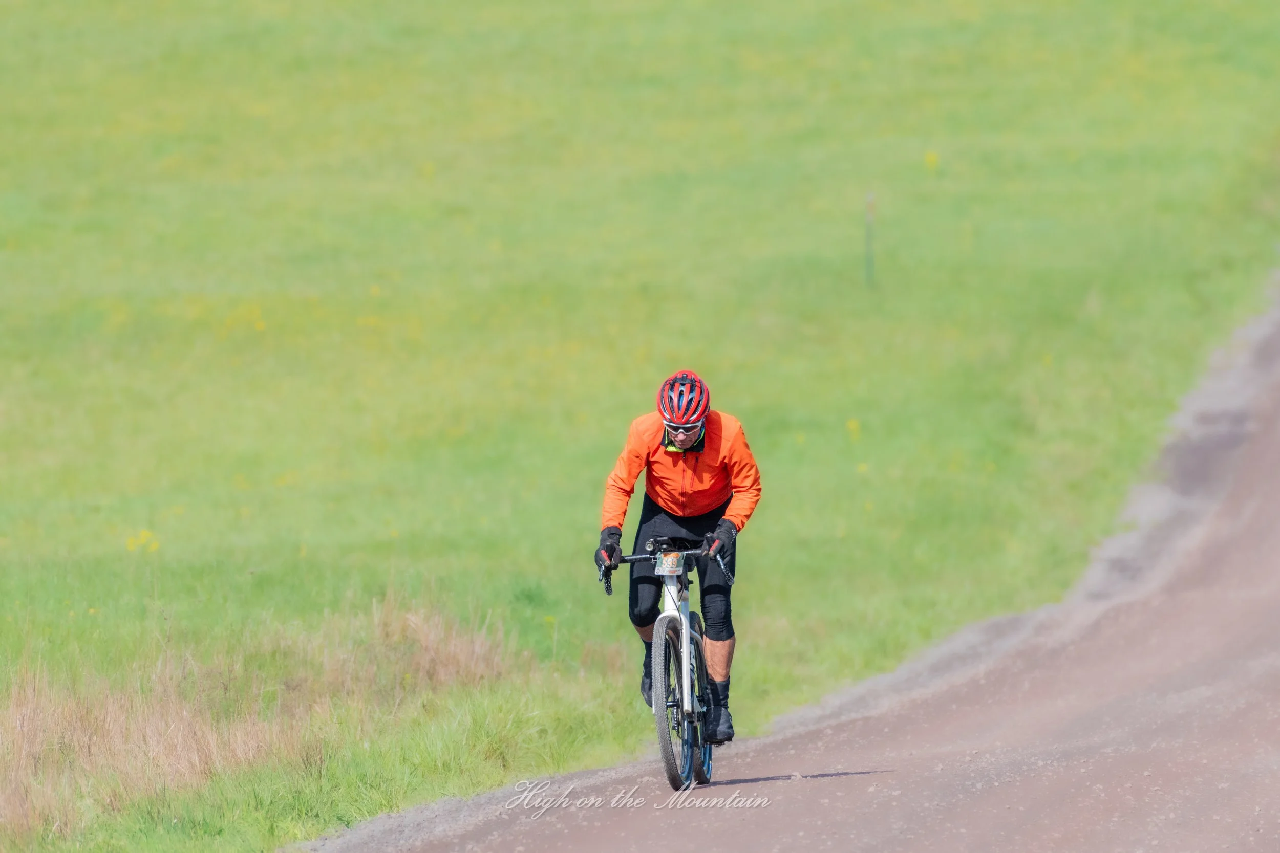 A person riding a mountain bike on a dirt track through a green grassy field, wearing a red helmet and orange jacket.