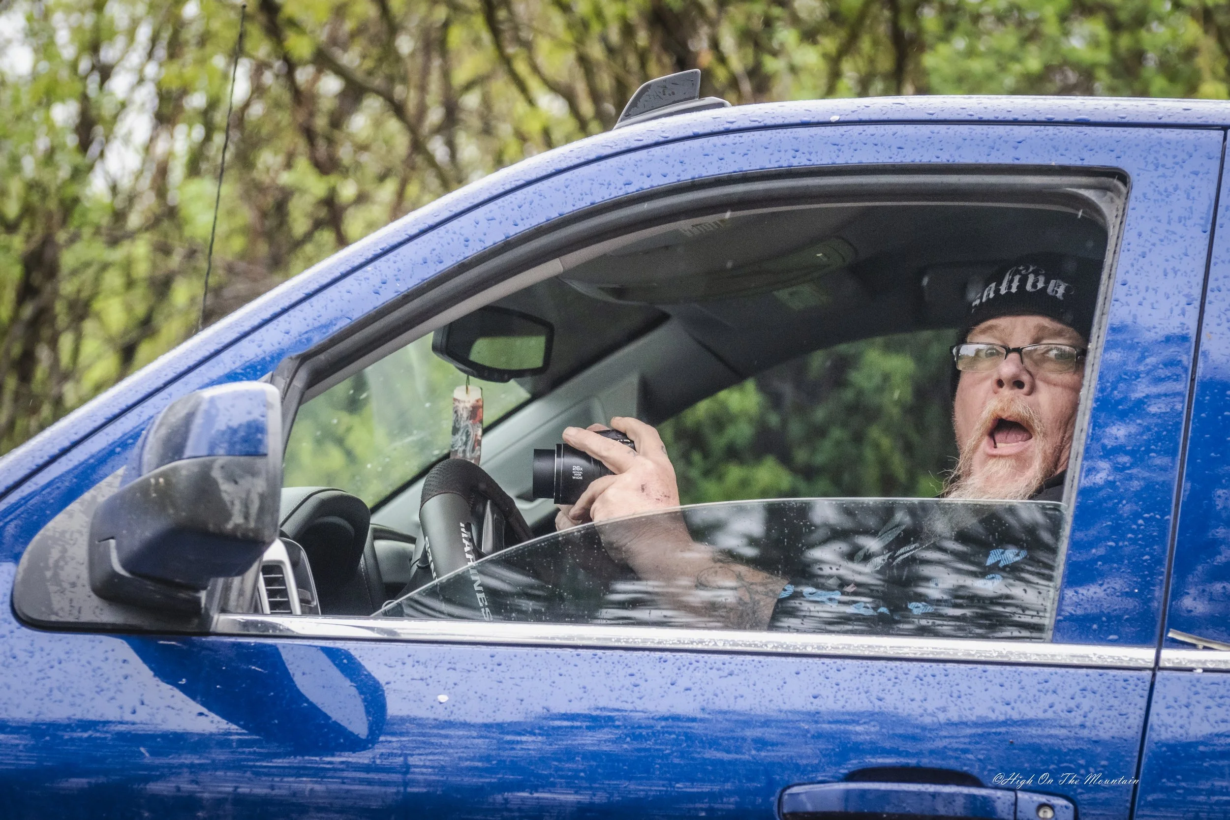 A man with glasses, a beard, and a black beanie sitting in a blue vehicle, holding a camera and looking out the window during rainy weather with trees in the background.