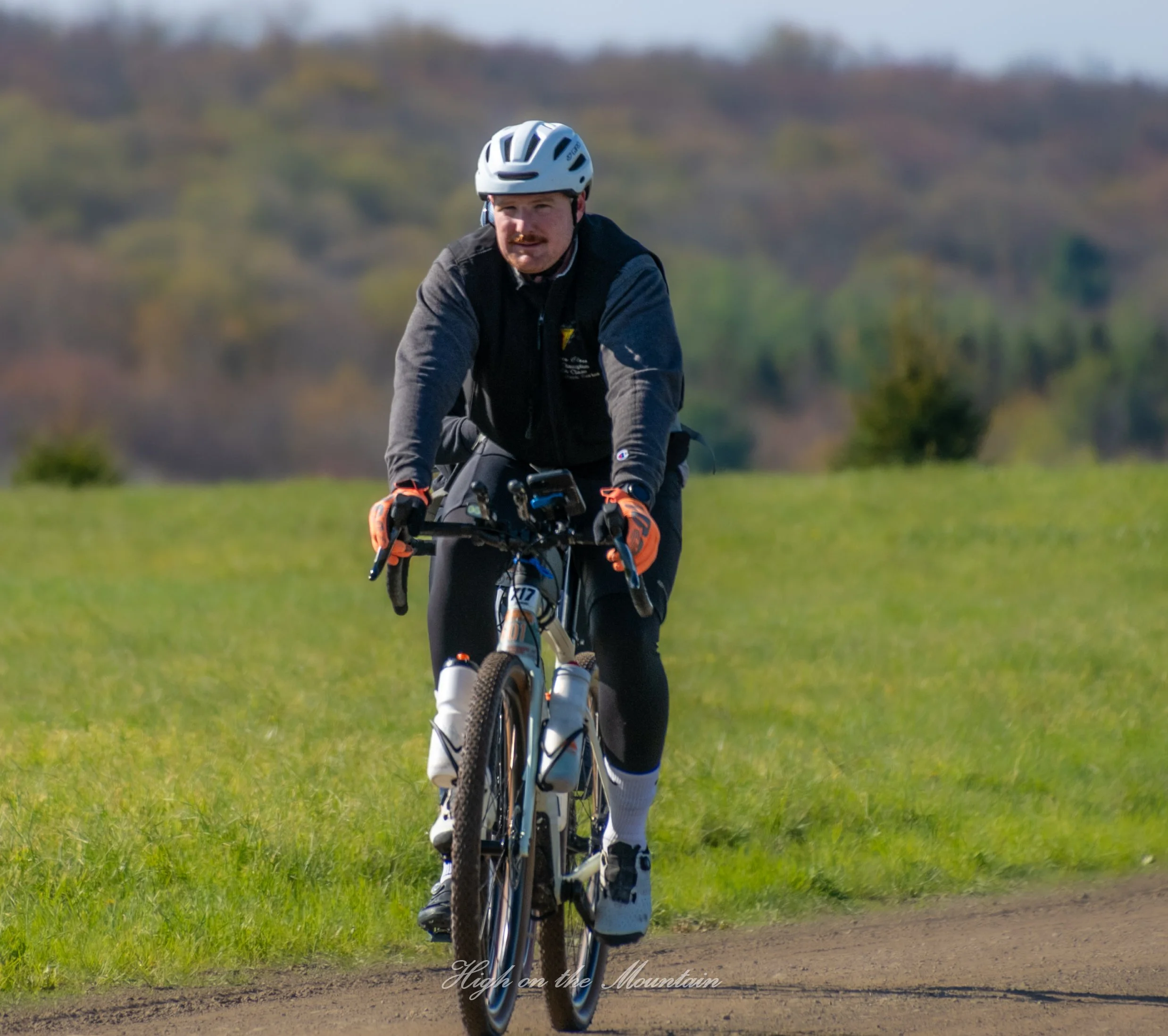 A man wearing a white helmet and black jacket riding a mountain bike on a grassy trail with trees and hills in the background.