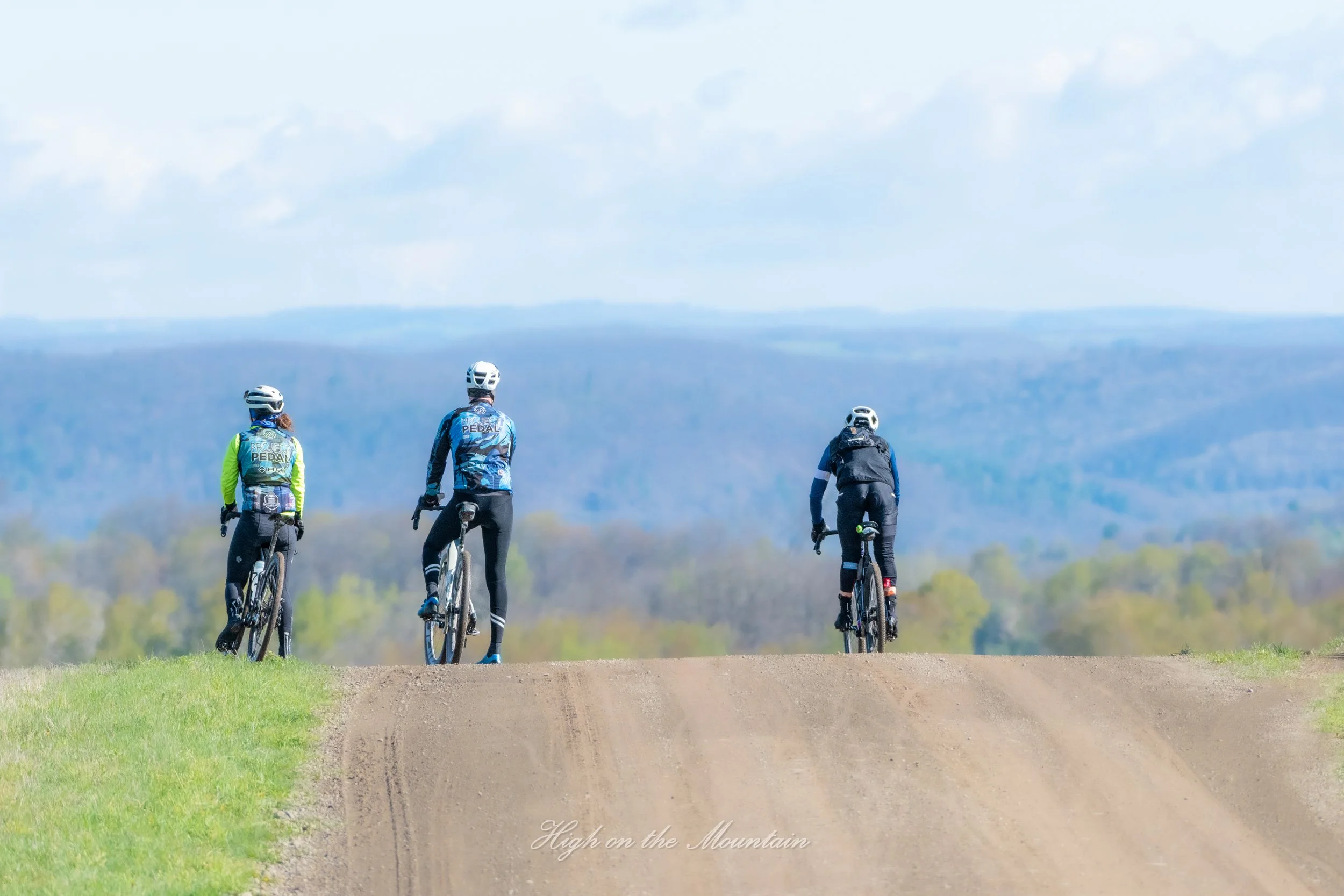 Four children riding bicycles on a dirt trail in a scenic outdoor area with mountains and sky in the background.