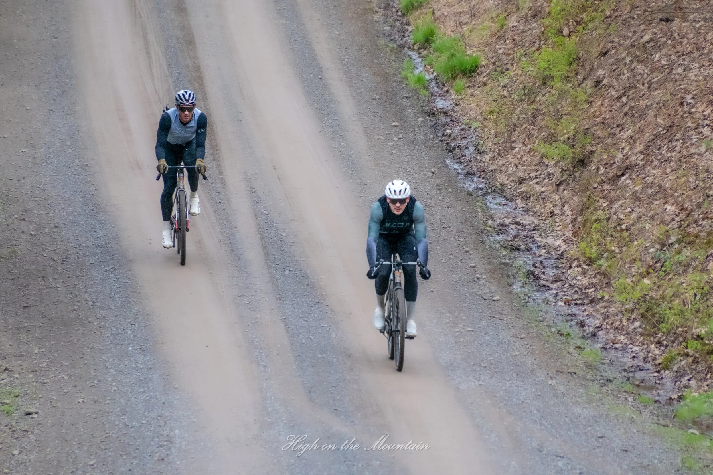 Two cyclists riding on a dirt mountain trail with green vegetation on the side.