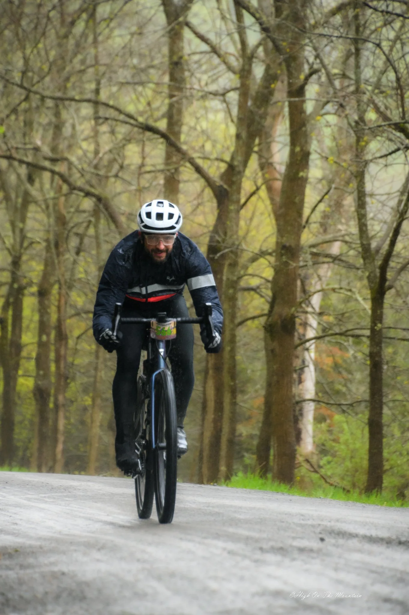 A man biking on a trail through a wooded area with tall trees and falling leaves, wearing a helmet, glasses, and biking gear.