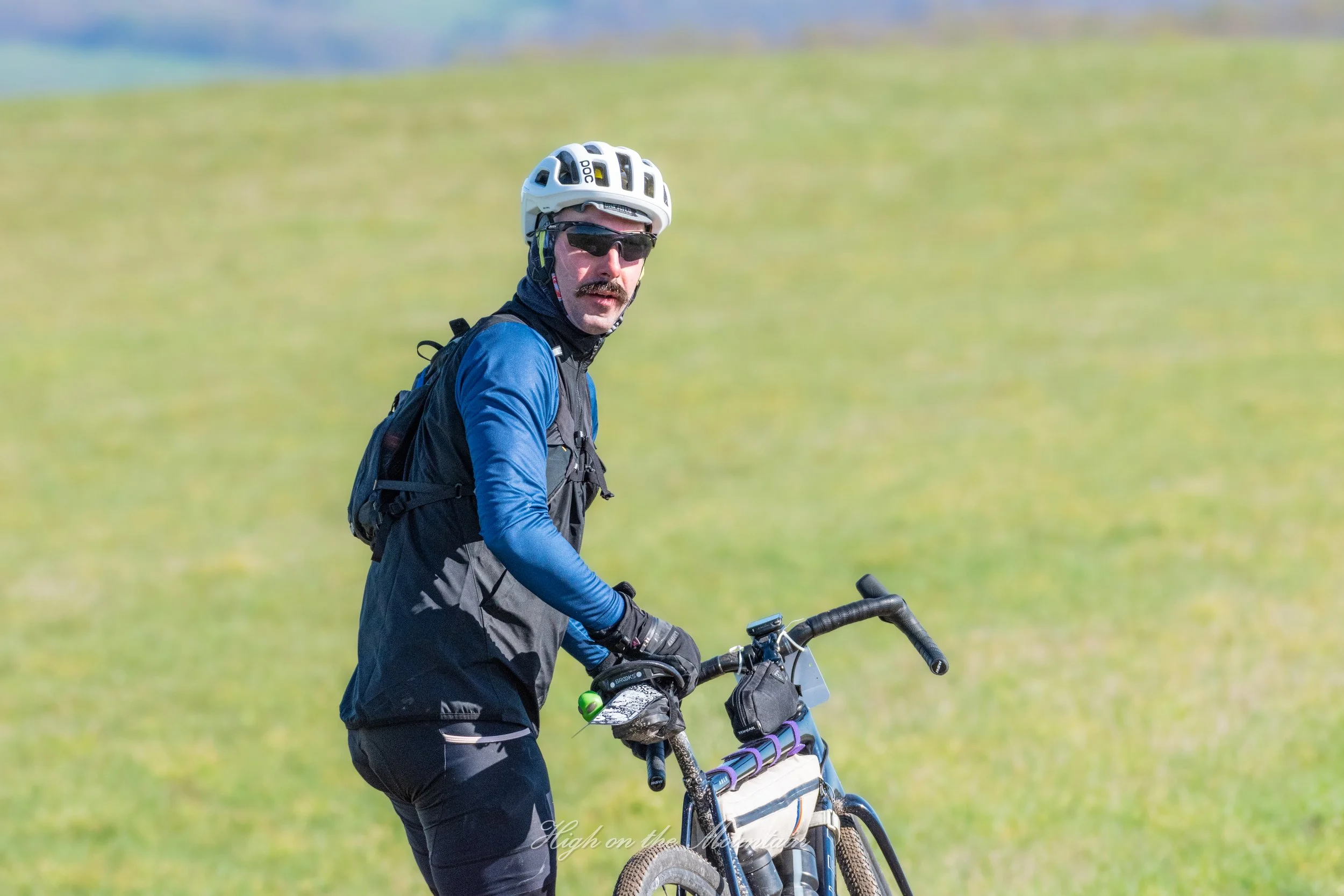 A man wearing a white helmet, sunglasses, a black vest, and blue long-sleeve shirt, riding a mountain bike in a grassy field.