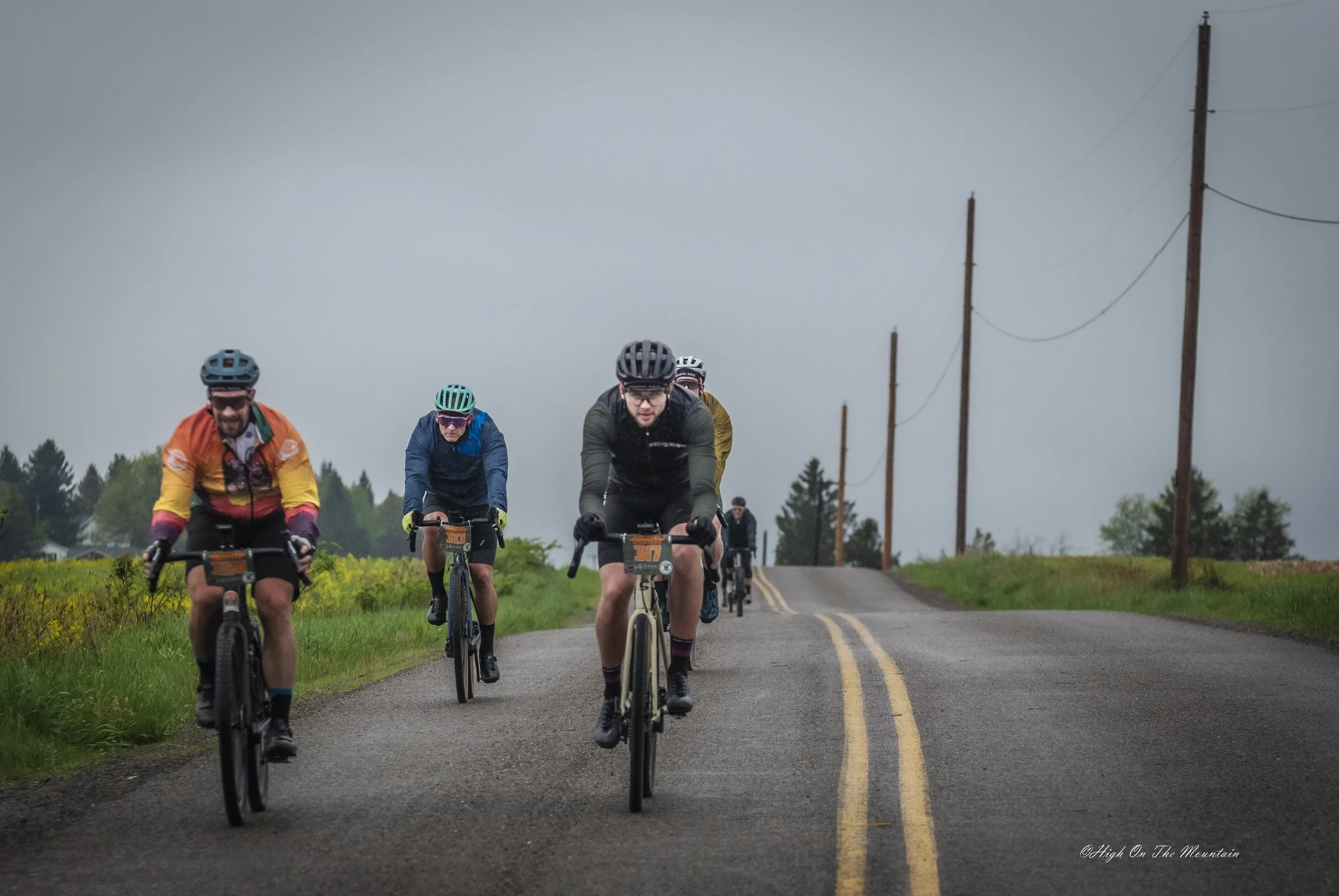 A group of five cyclists riding on a rural road under a cloudy sky.
