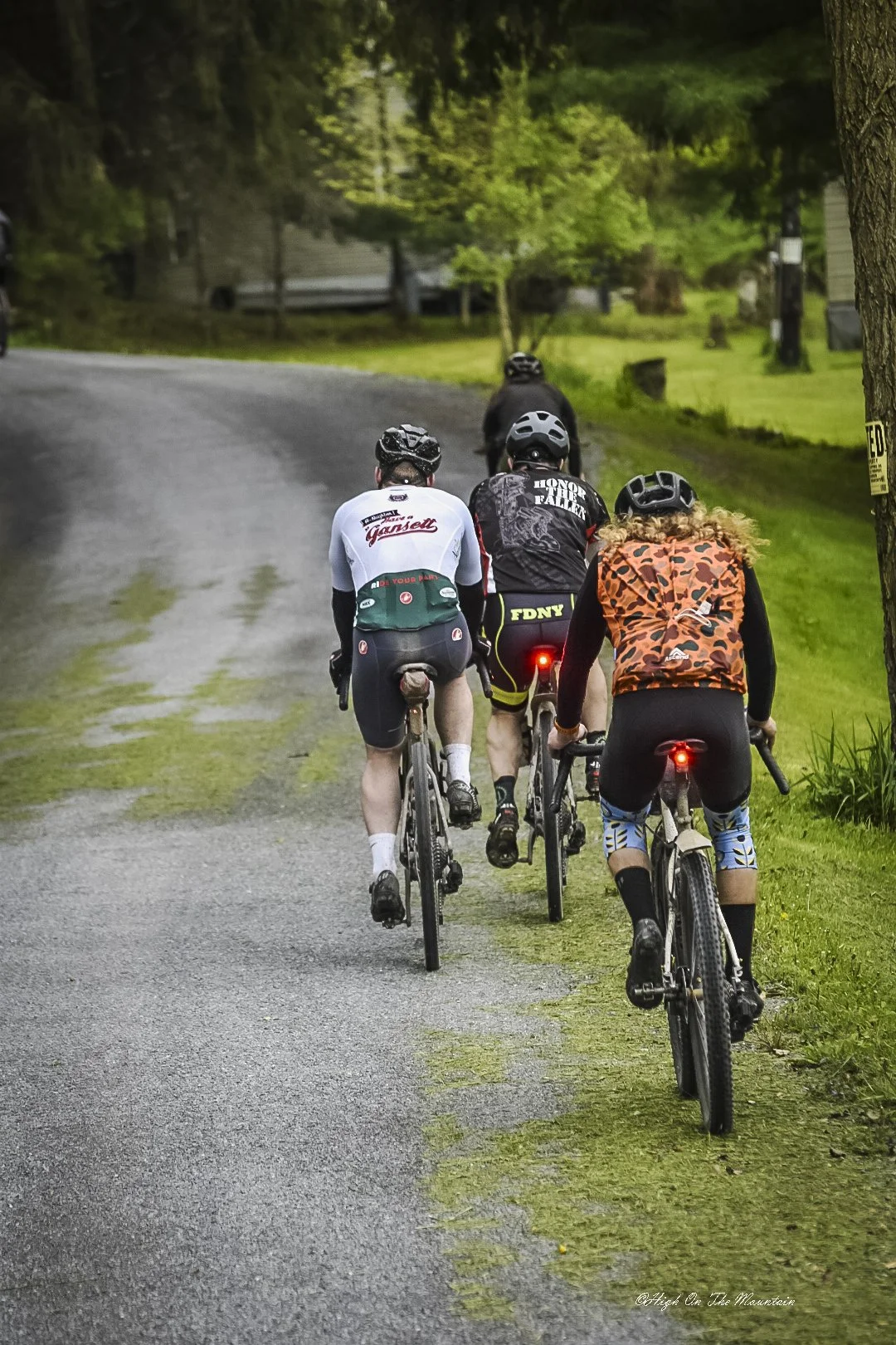 Four cyclists riding on a gravel trail through a green, forested area, viewed from behind.