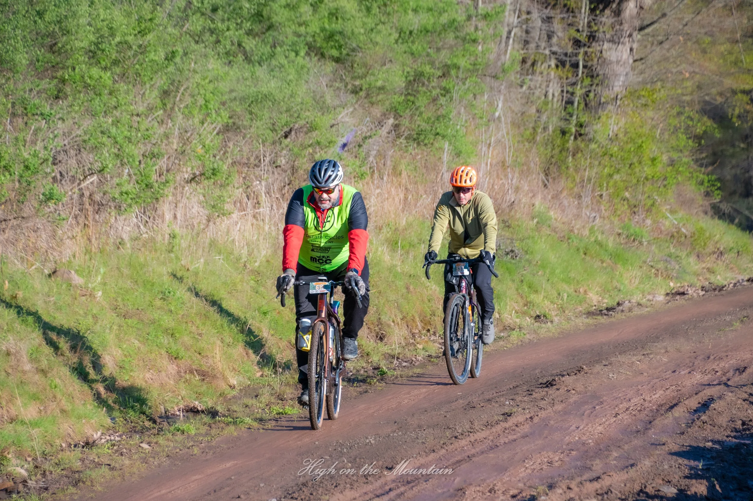 Two cyclists riding mountain bikes on a dirt trail through green and brown foliage on a sunny day.