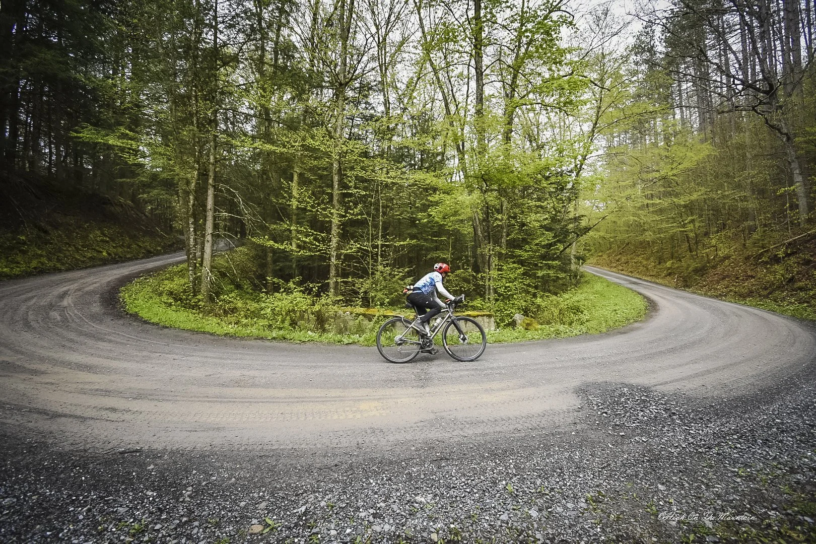 A cyclist wearing a red helmet and blue jacket riding a mountain bike on a dirt switchback road through a dense green forest.