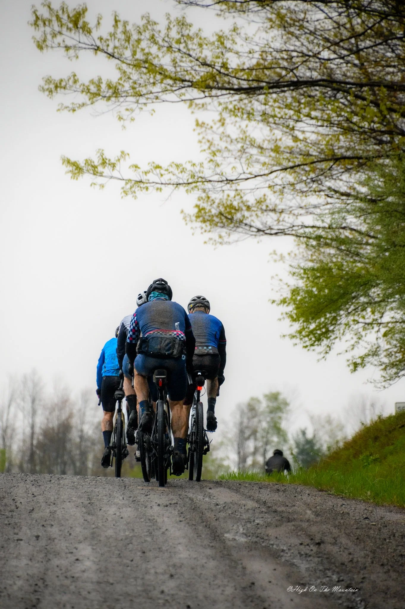 A group of cyclists riding on a dirt path surrounded by trees with green leaves, with a cloudy sky overhead.