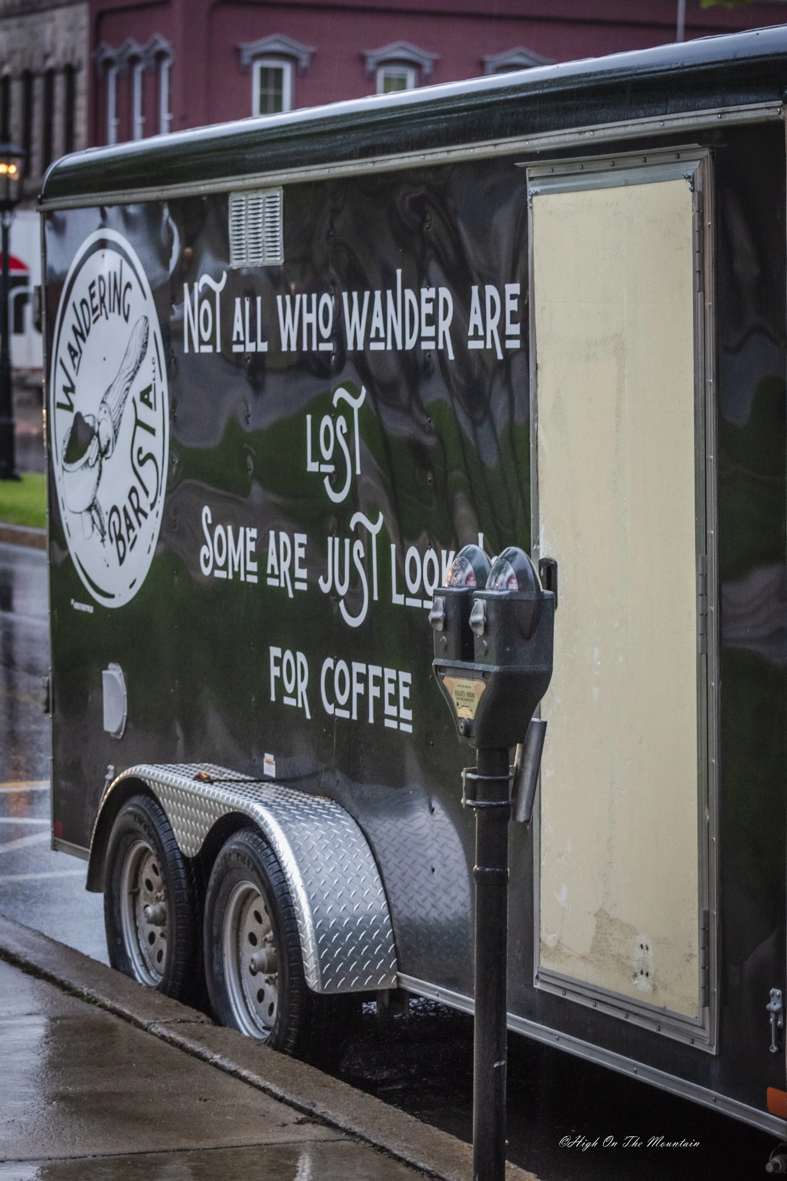 A black food trailer with white text and a logo parked on a wet street at dusk.