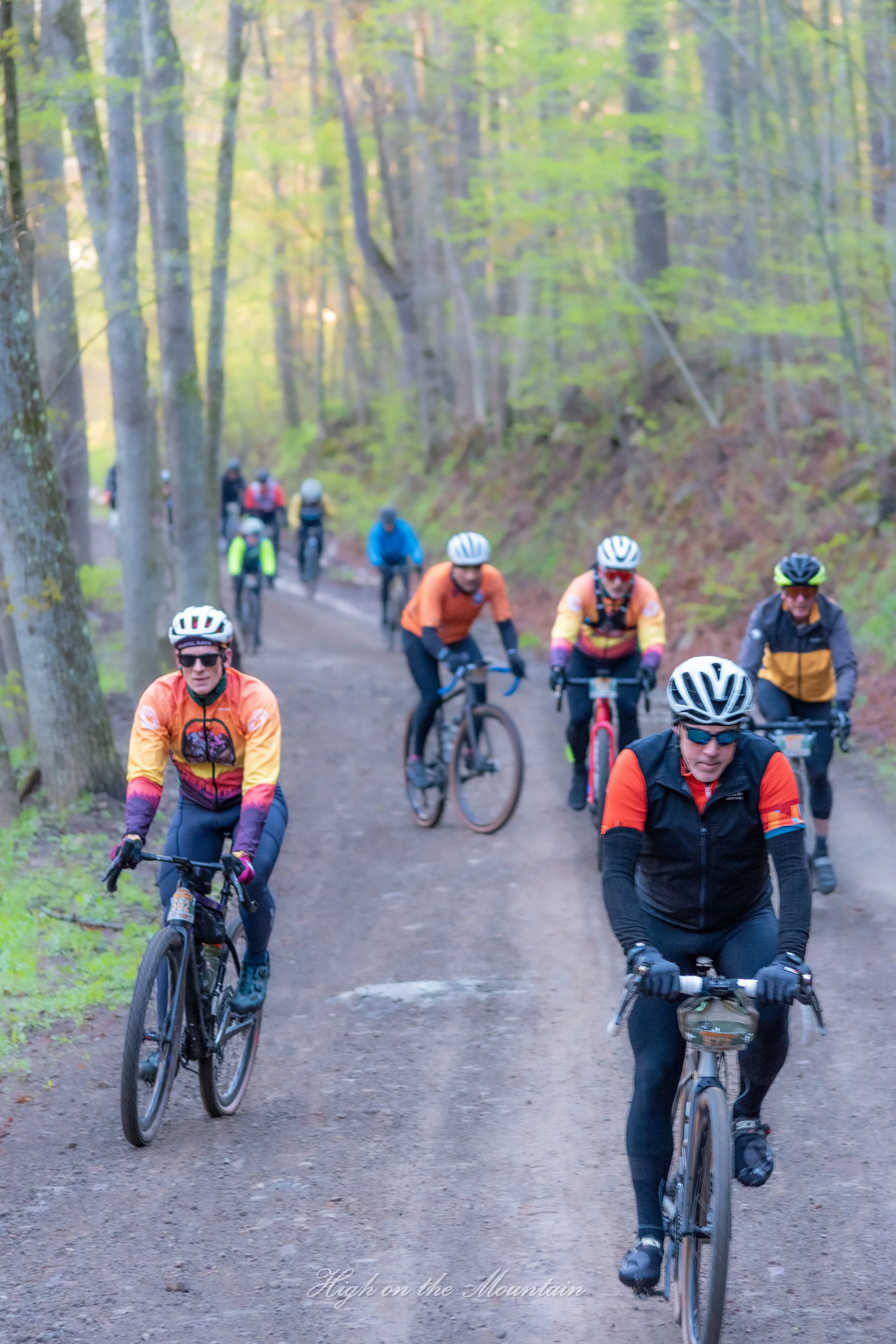 A group of mountain bikers riding on a dirt trail through a forested area with tall trees and green foliage. The bikers are wearing helmets and colorful outdoor gear.