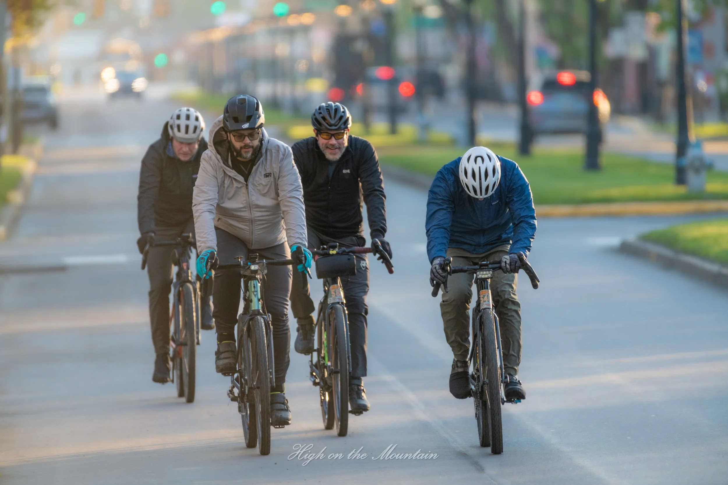 Four men riding bikes together on a street during early morning or late afternoon, wearing helmets and jackets, with blurred cars and trees in the background, and the words 'High on the Mountain' at the bottom.