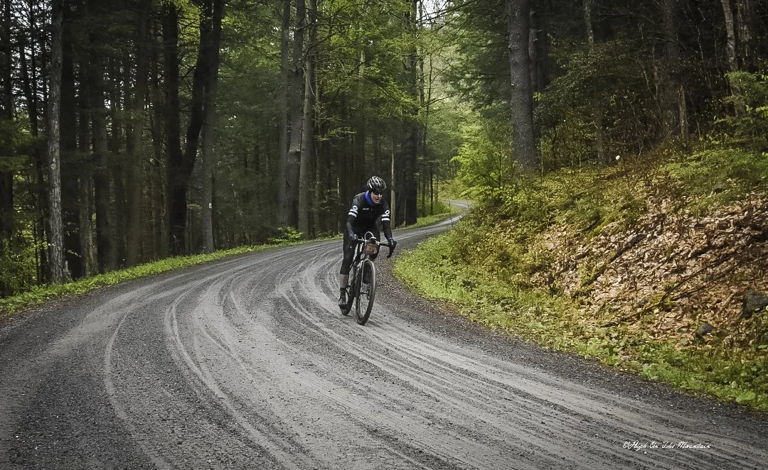 A cyclist riding on a winding gravel forest trail surrounded by tall trees and green foliage.