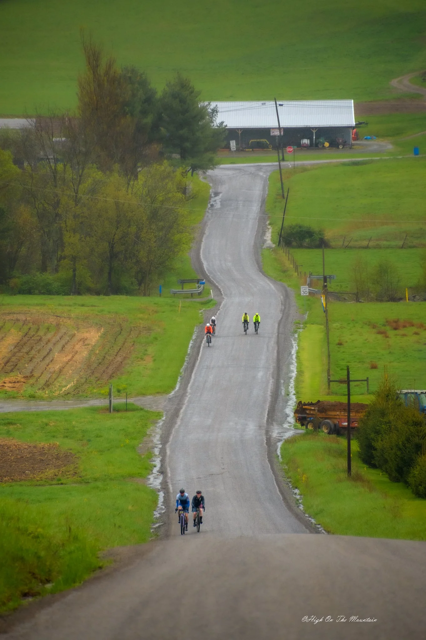 A rural scene with a long gravel road winding through green fields and trees. Several groups of cyclists are riding along the road. In the background, there is a large barn and a few vehicles. Utility poles line the road, and the sky is overcast.
