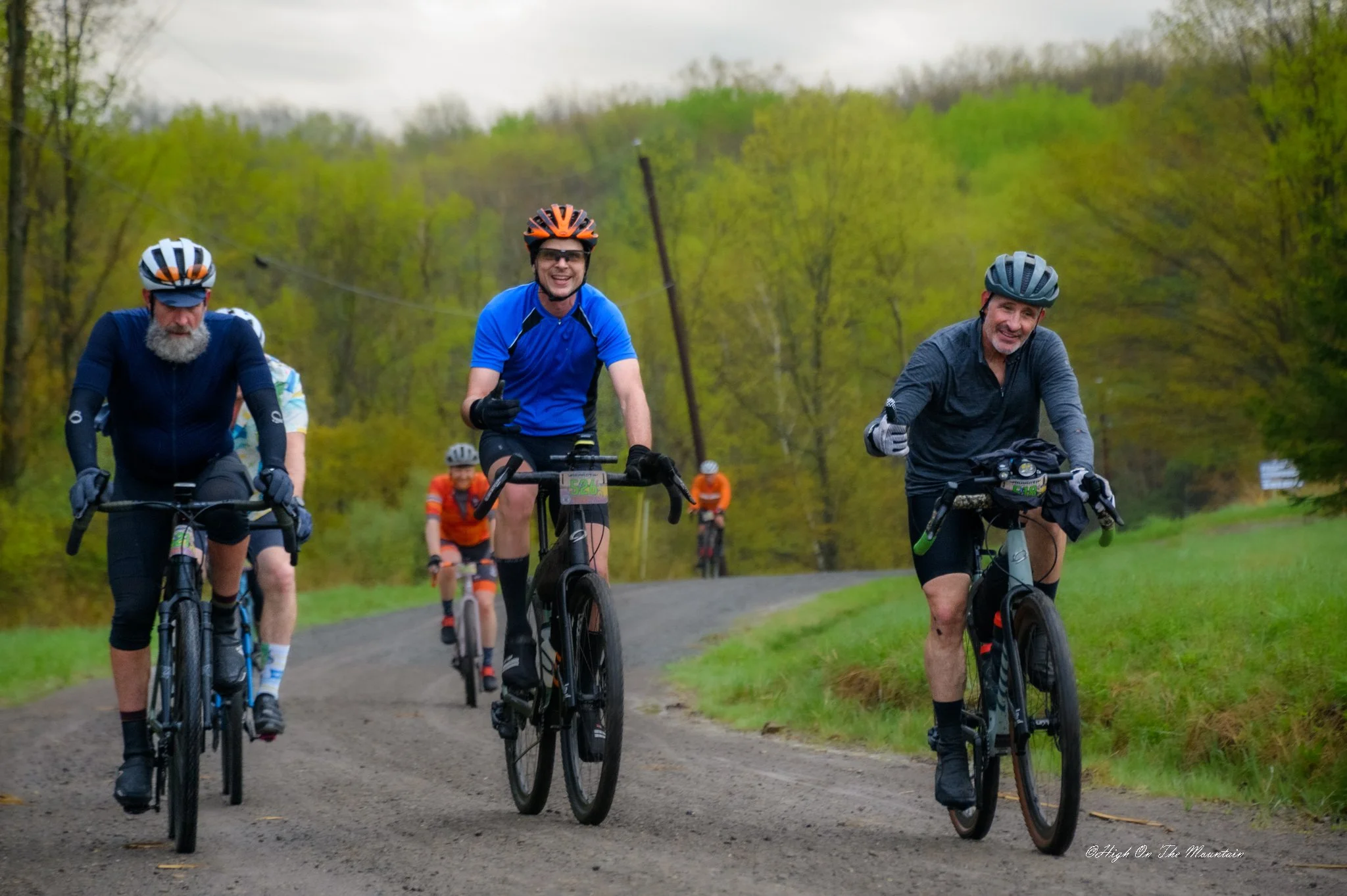 Group of men riding bicycles on a dirt trail through a green, wooded area on a cloudy day, smiling and enjoying the ride.