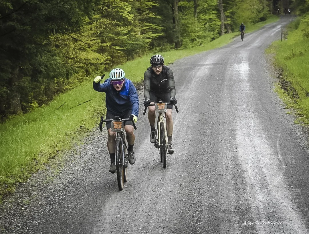 Two cyclists riding on a gravel road through a green, wooded area, with one cyclist giving a thumbs up and the other looking ahead.