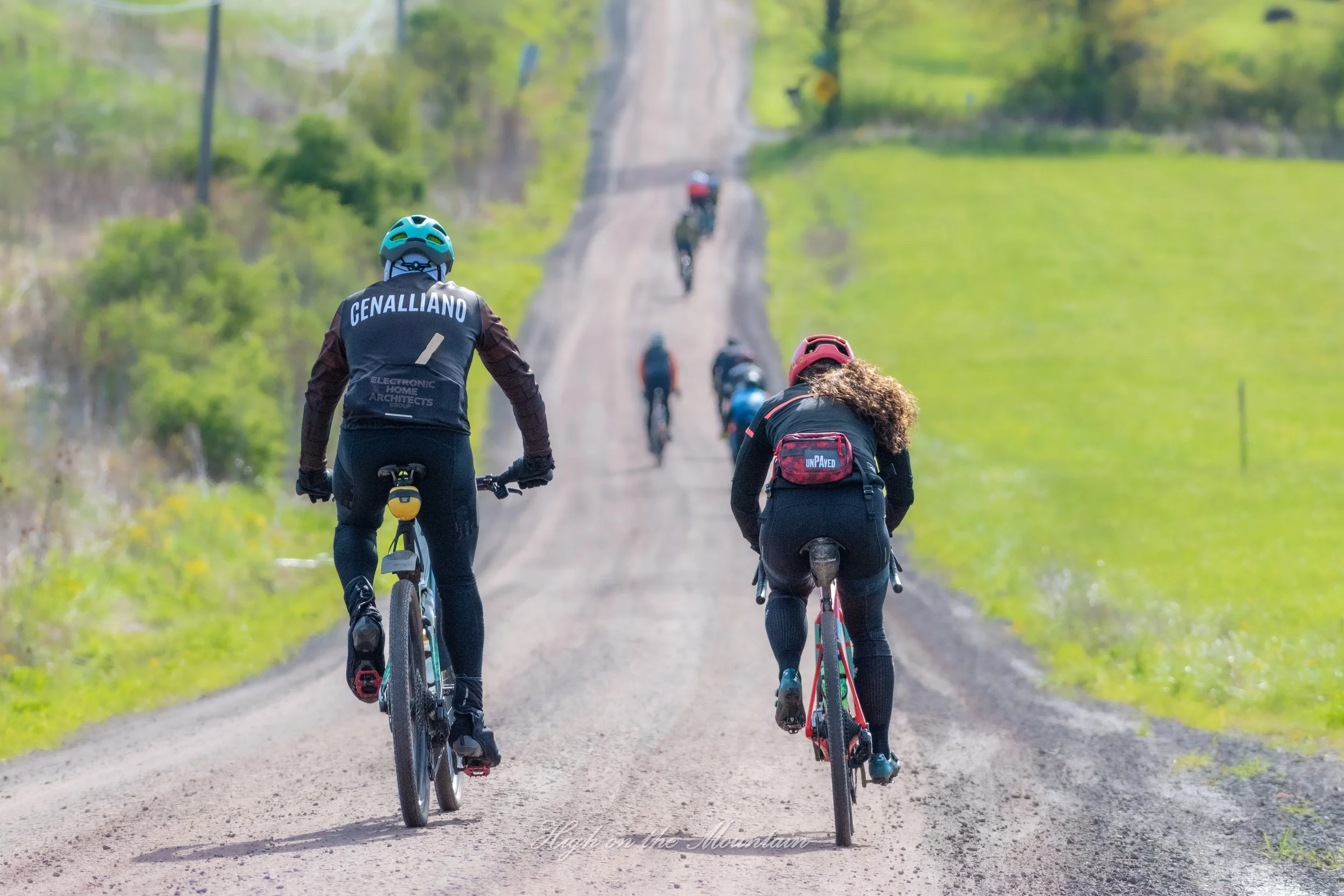 Two women riding bikes on a dirt trail through a green, rural landscape with other cyclists ahead of them.
