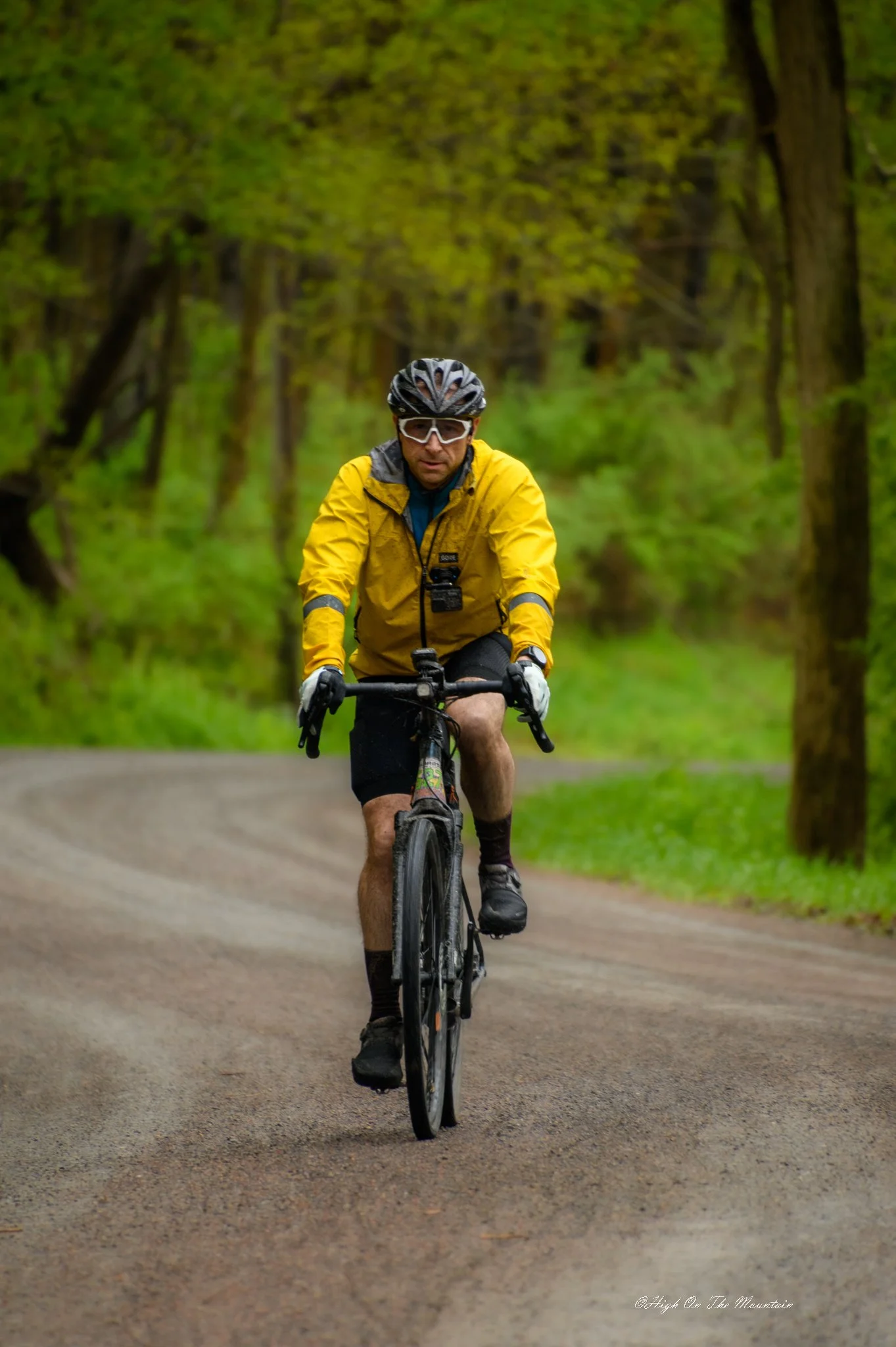 A man riding a mountain bike on a dirt path through a green forest. He is wearing a yellow jacket, black helmet, and sports glasses.