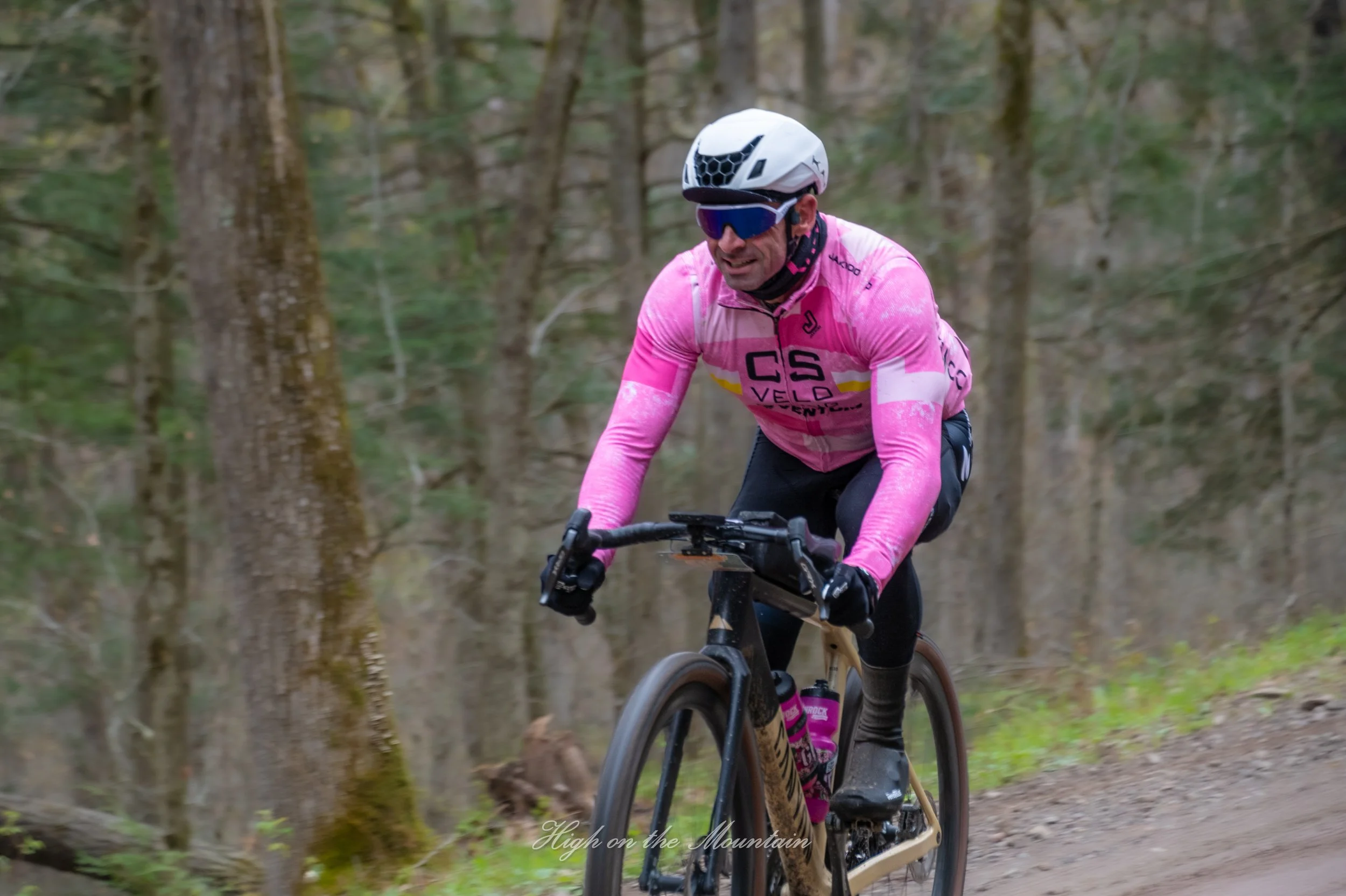 A man wearing a pink cycling jersey, black helmet, and sunglasses riding a mountain bike through a forest trail surrounded by trees.