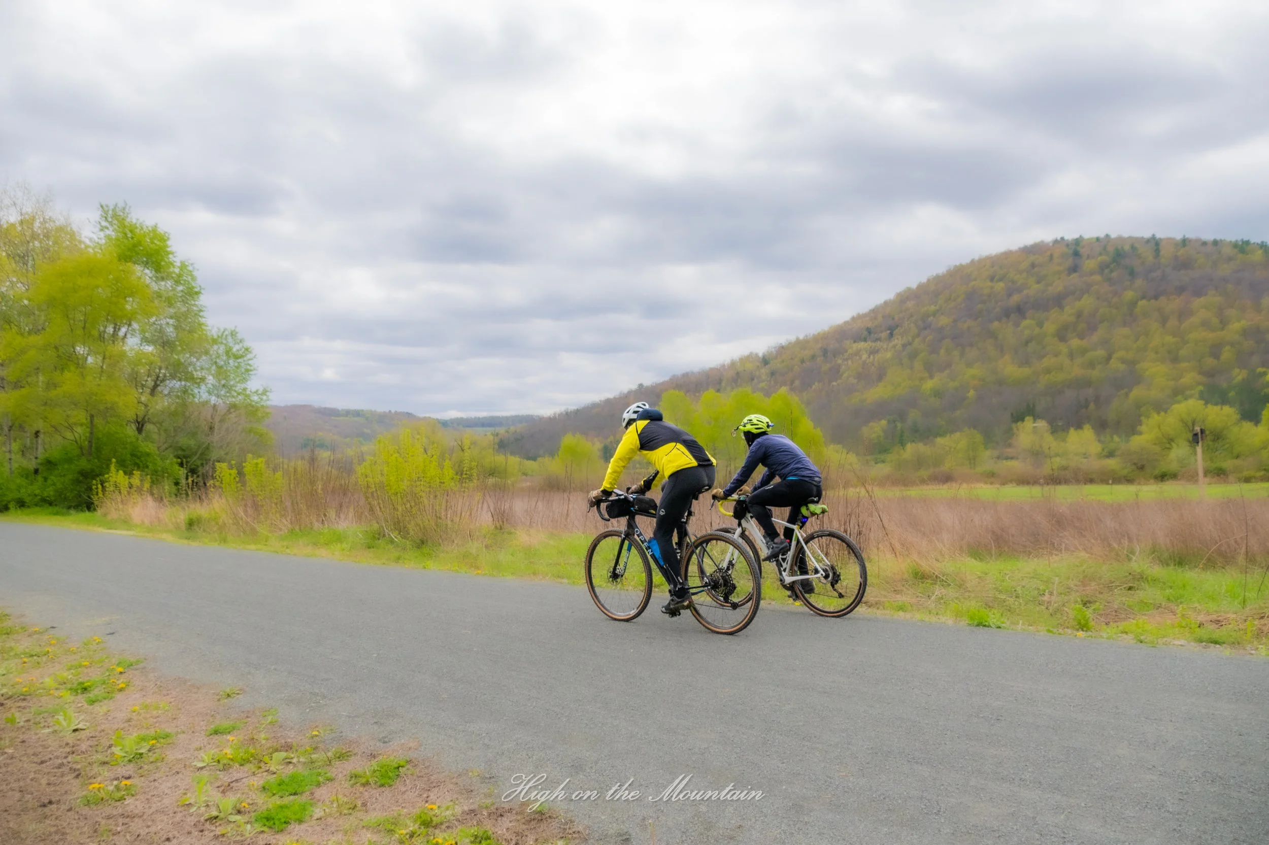 Two people riding bicycles on a paved path in a scenic mountain landscape with green trees and overcast sky.